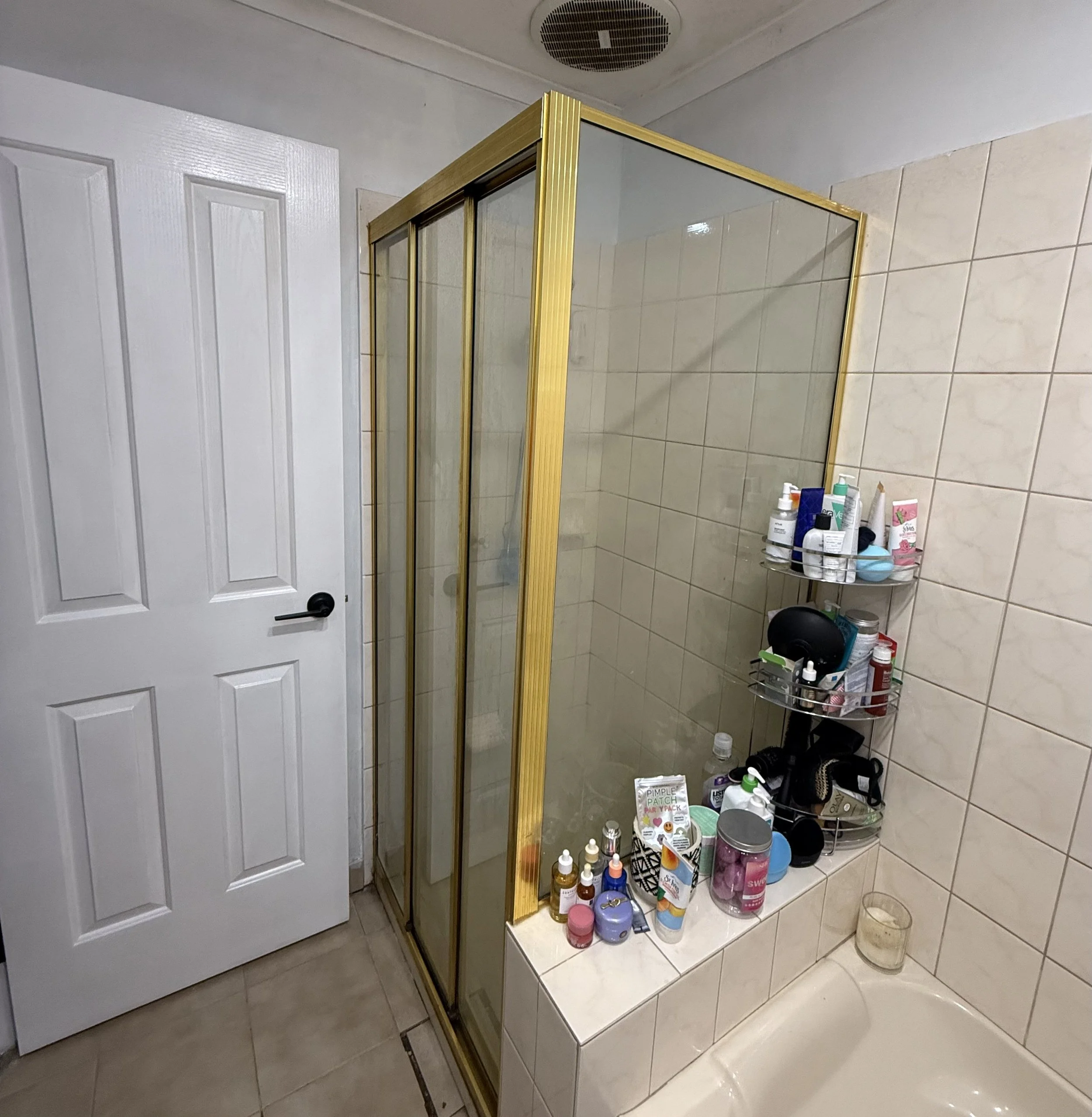 A bathroom with a white door, a gold-framed glass shower enclosure, and a small shelf with various toiletries on a tiled wall and floor.