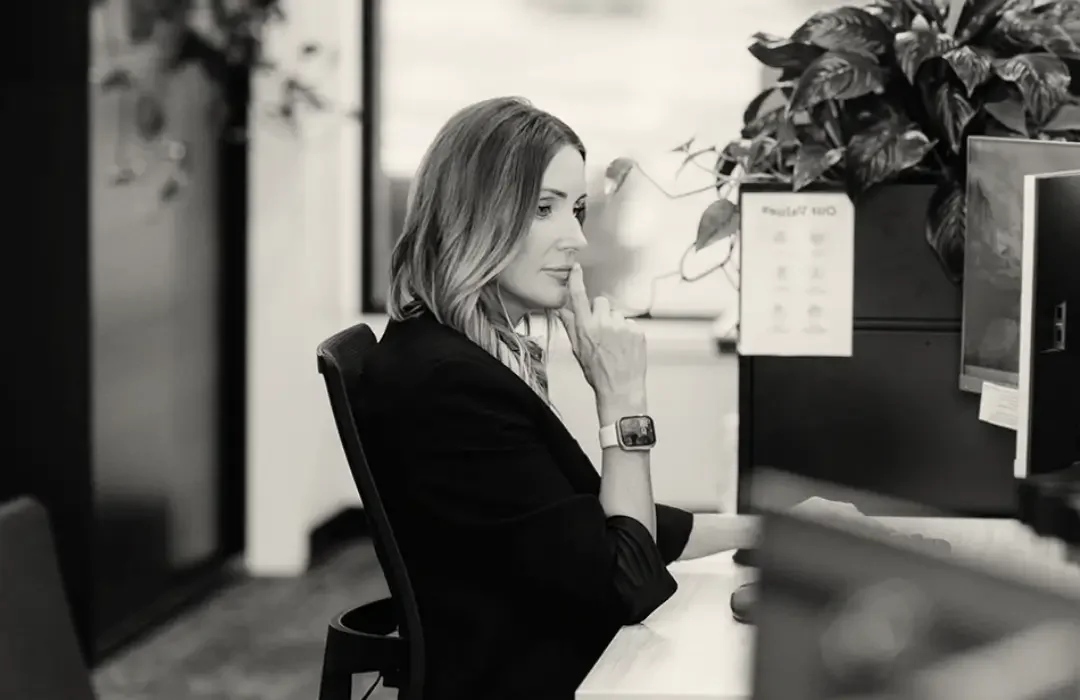 A woman sitting at a desk in an office, wearing a smartwatch and a blazer, with her hand on her chin, looking at her computer screen.