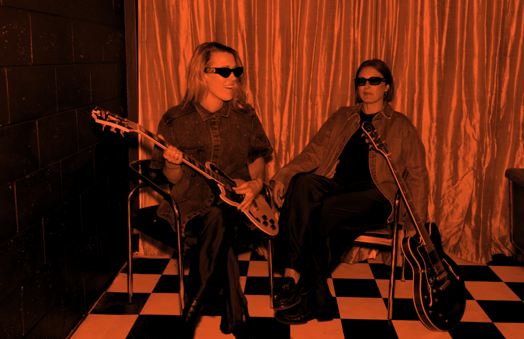 Assembl founders Jade Burger and Nat Eady sitting on chairs in a room with checkered floor, playing guitars, wearing sunglasses and casual clothing, with a curtain in the background.