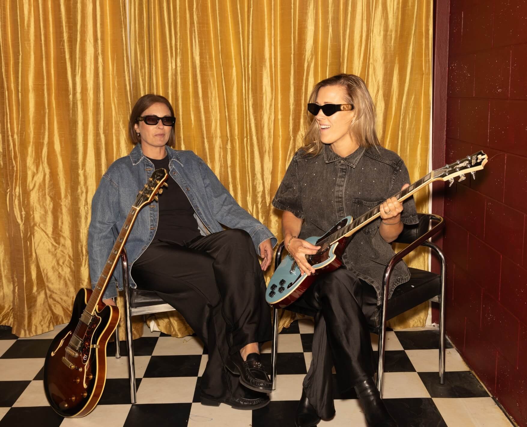Two women with guitars sitting in a room with a checkered floor, gold curtain, and red wall, wearing black sunglasses and dark clothing.