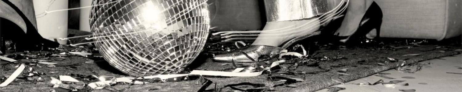 Close-up editorial image of a disco ball, metallic heels and scattered party streamers on the floor in warm monochrome lighting, used as the Assembl Studio website footer image.