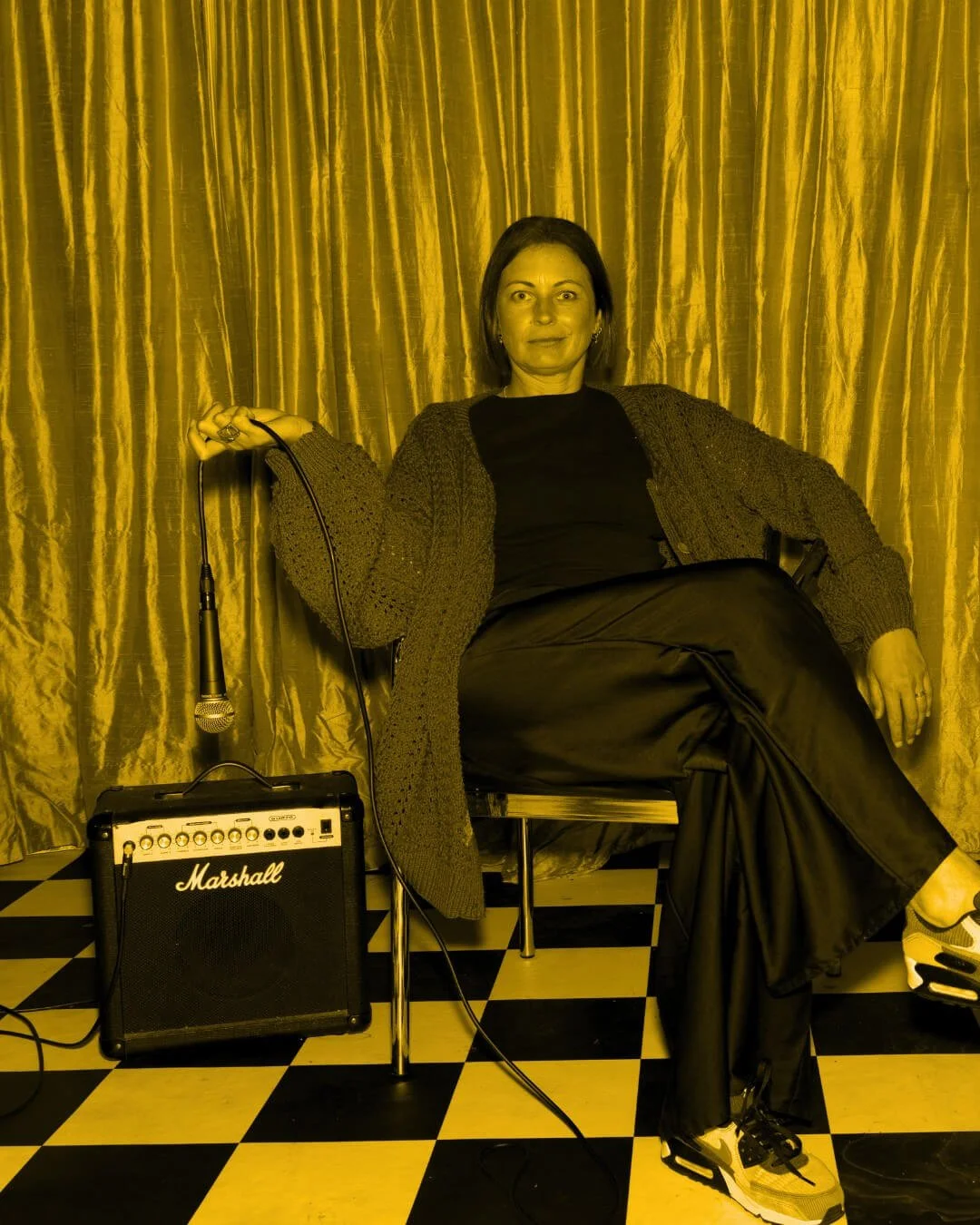Portrait of Jade Burger, co-founder of Assembl Studio, seated with a microphone and Marshall amplifier against gold stage curtains and a black-and-white checkered floor, styled in a bold editorial yellow monochrome.
