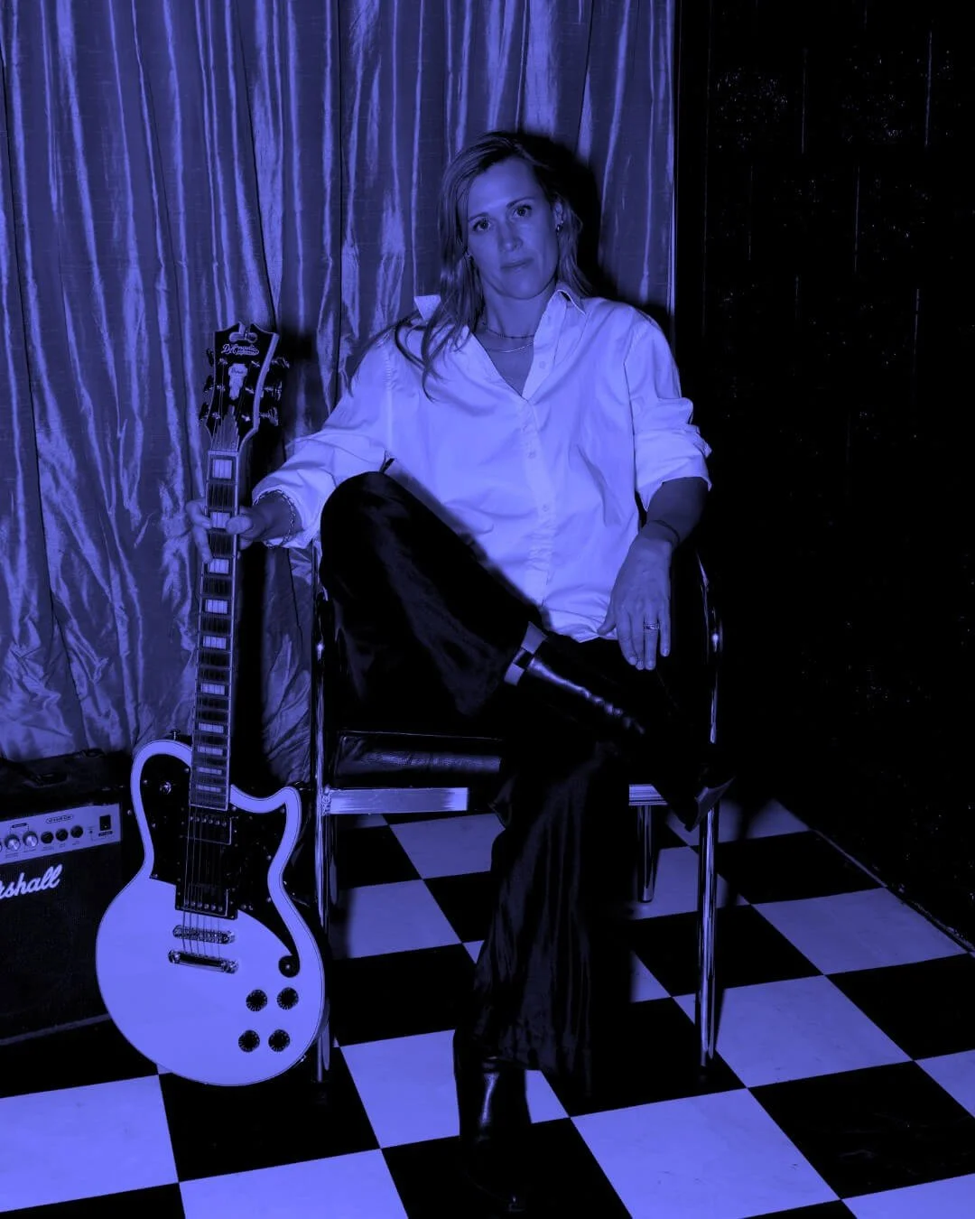 Portrait of Nat Eady, co-founder of Assembl Studio, seated beside a white electric guitar against velvet curtains and a checkered floor, styled in a bold editorial blue monochrome.