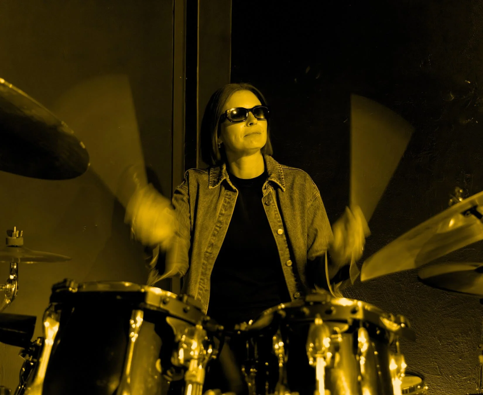 Jade Burger, Assembl Studio founder playing drums in a dark studio setting, wearing sunglasses and a denim jacket.