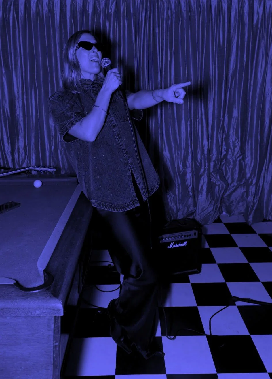 Editorial portrait of Nat Eady, co-founder of Assembl Studio, singing into a microphone beside a pool table and Marshall amplifier in dramatic blue monochrome lighting.