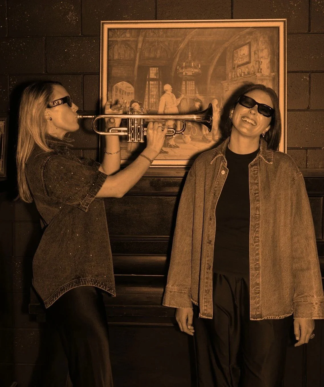 Assembl founders Jade Burger and Nat Eady wearing dark sunglasses, one playing a trumpet and the other smiling, standing in front of a framed painting on a dark wall.