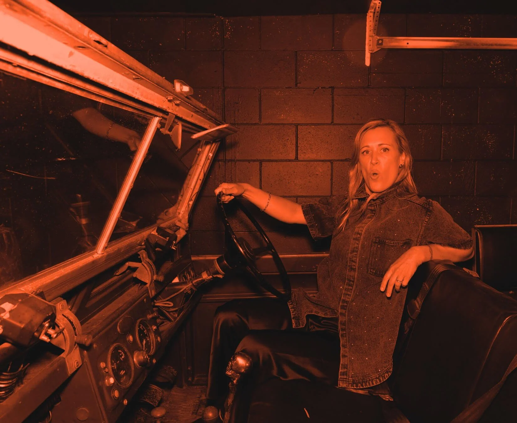 Editorial portrait of Nat Eady, co-founder of Assembl Studio, seated inside a vintage vehicle in a garage setting with dramatic red monochrome lighting.
