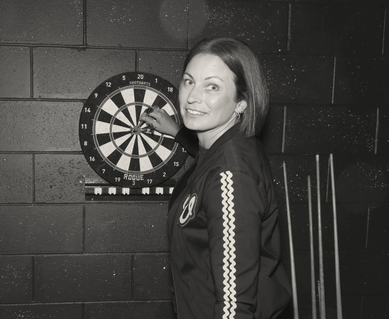 Black-and-white portrait of Jade Burger, co-founder of Assembl Studio, standing beside a dartboard in a moody games-room setting with pool cues and textured black walls.