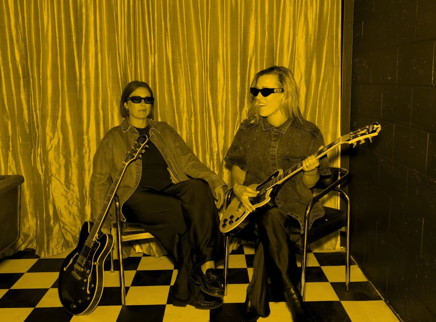 Two assembl founders sitting on chairs with guitars; one woman has a black guitar on the floor, the other is holding a white electric guitar; both wearing sunglasses and denim jackets, sitting in front of a gold curtain and black-studded wall.