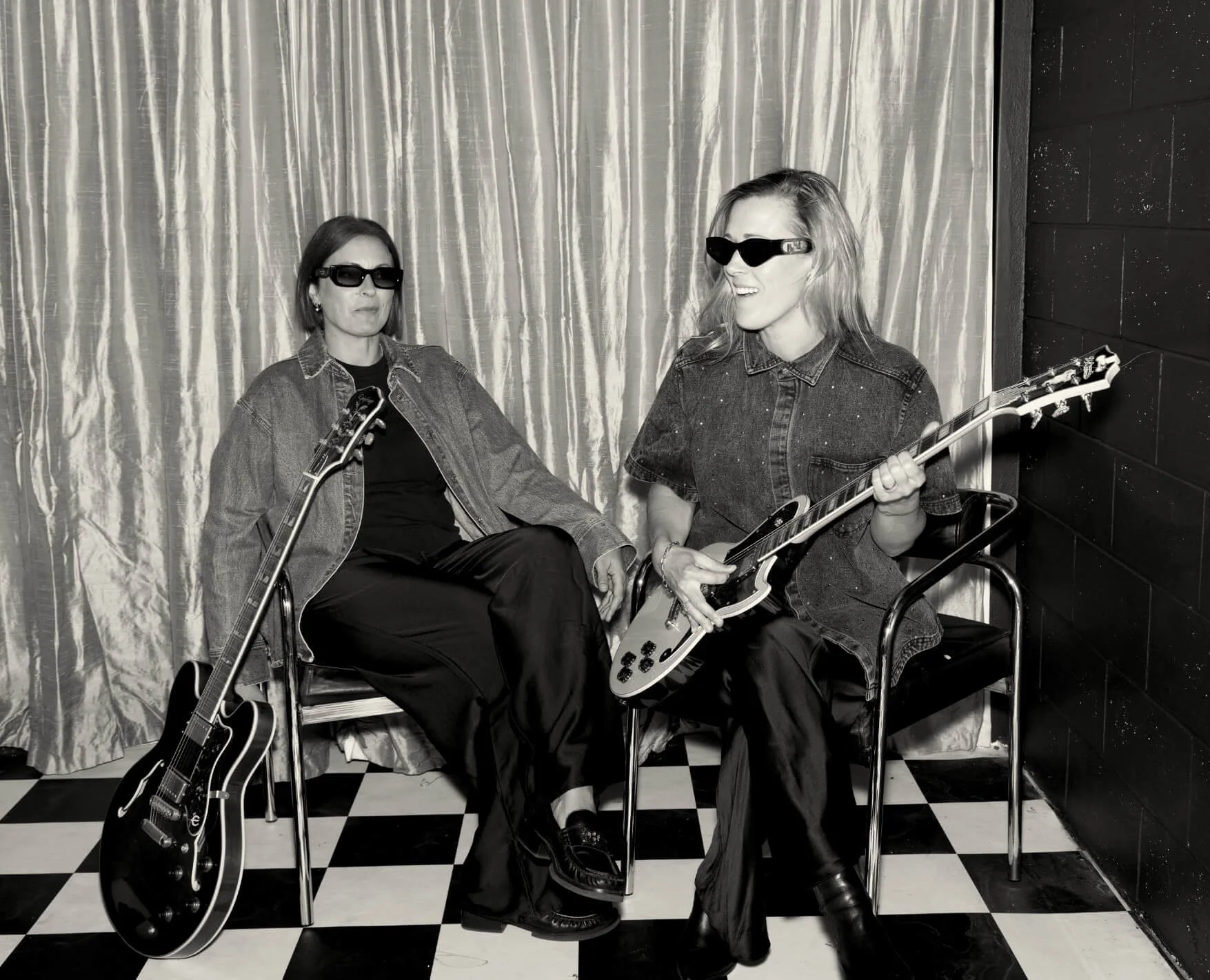 Black-and-white editorial portrait of Assembl Studio co-founders Nat Eady and Jade Burger seated with electric guitars in a retro music-inspired setting with velvet curtains and a black-and-white checkered floor.