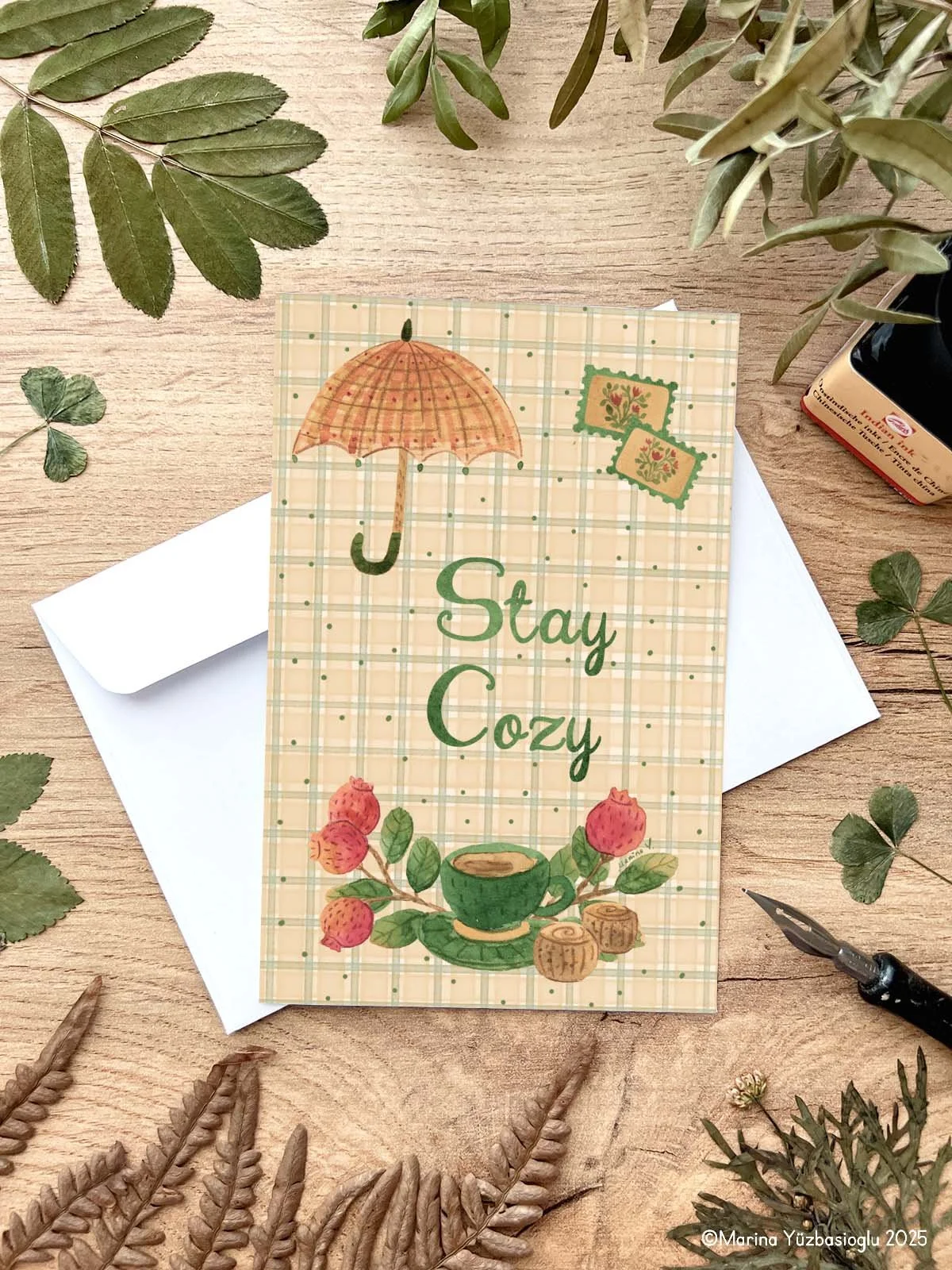 Featuring charming seasonal details—an umbrella, steaming teacup, rose hips, and hand-lettered text—this card captures the comforting spirit of autumn and winter.