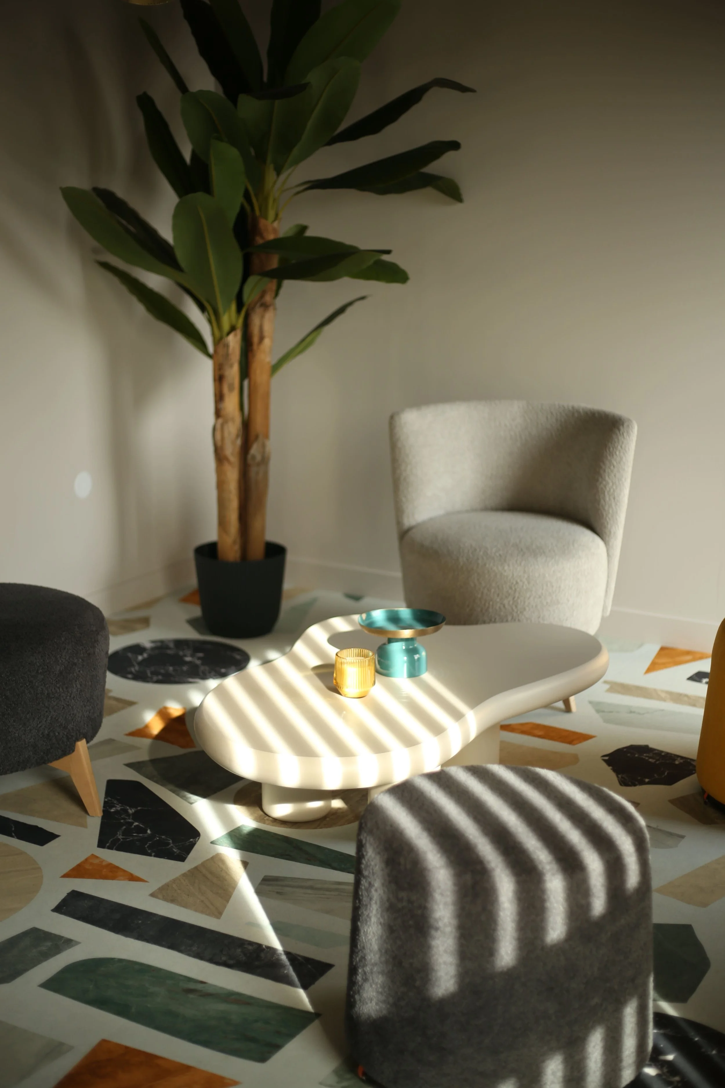 Salon intérieur avec un fauteuil beige, un tapis coloré, une table basse blanche, une grande plante verte et des objets décoratifs en verre jaune et turquoise, avec des rayures de lumière passant par des stores.