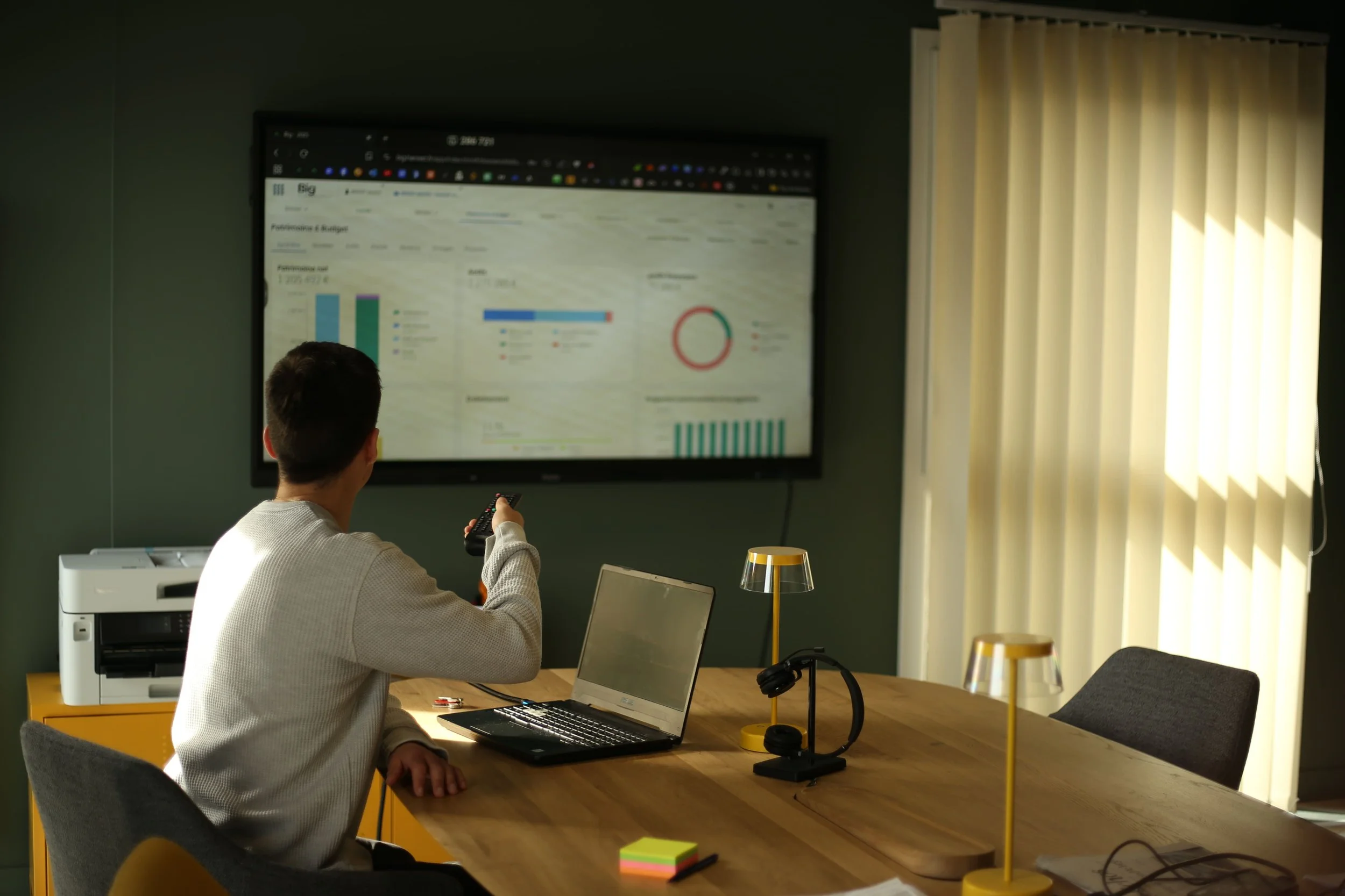 Un homme assis à une table d'entreprise regarde un écran d'ordinateur affichant des graphiques et des données, avec un télécommande à la main, dans un bureau éclairé par la lumière naturelle