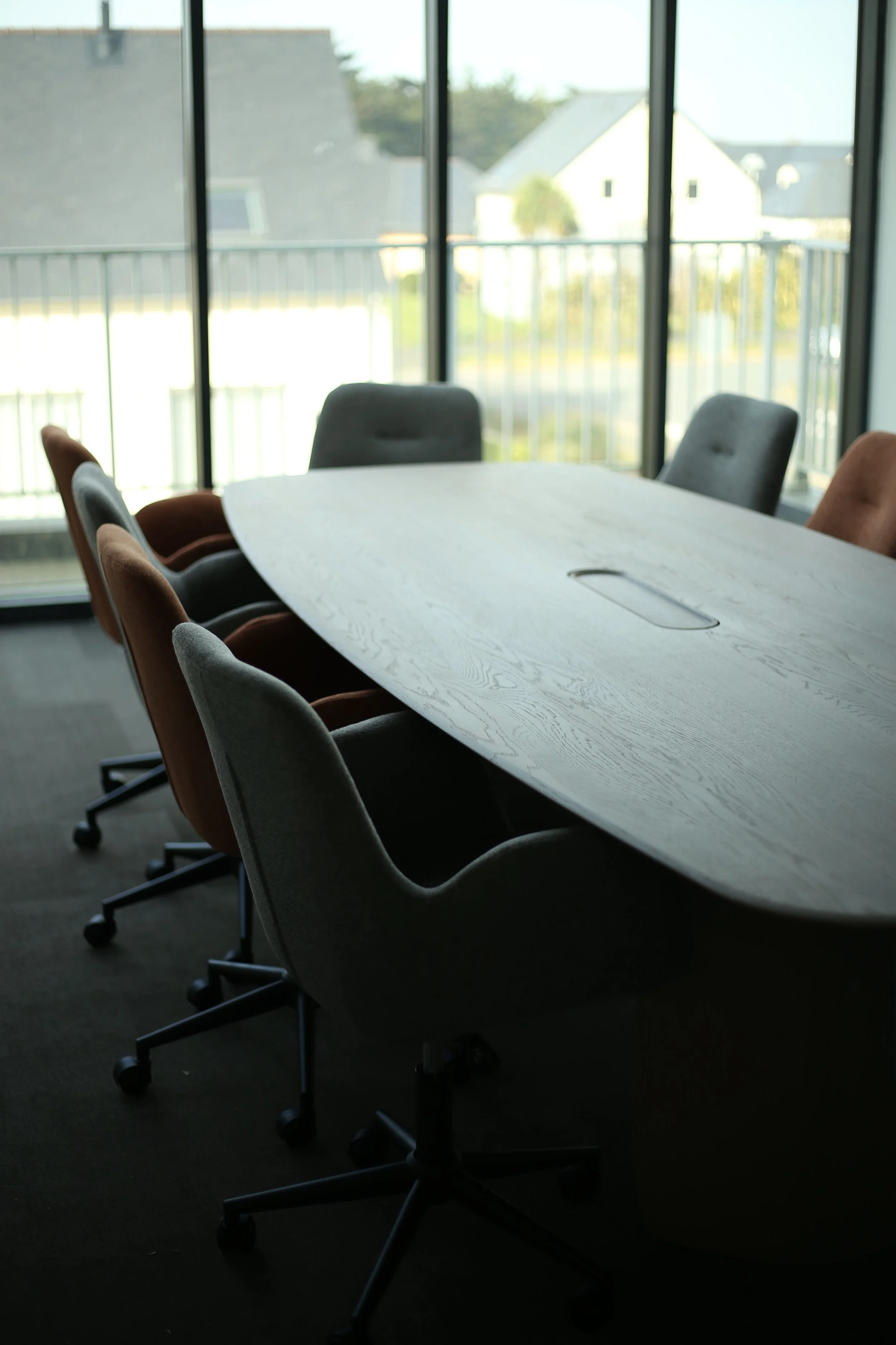 Salle de réunion avec une grande table en bois et plusieurs chaises de différentes couleurs autour, près de fenêtres donnant sur une terrasse extérieure.