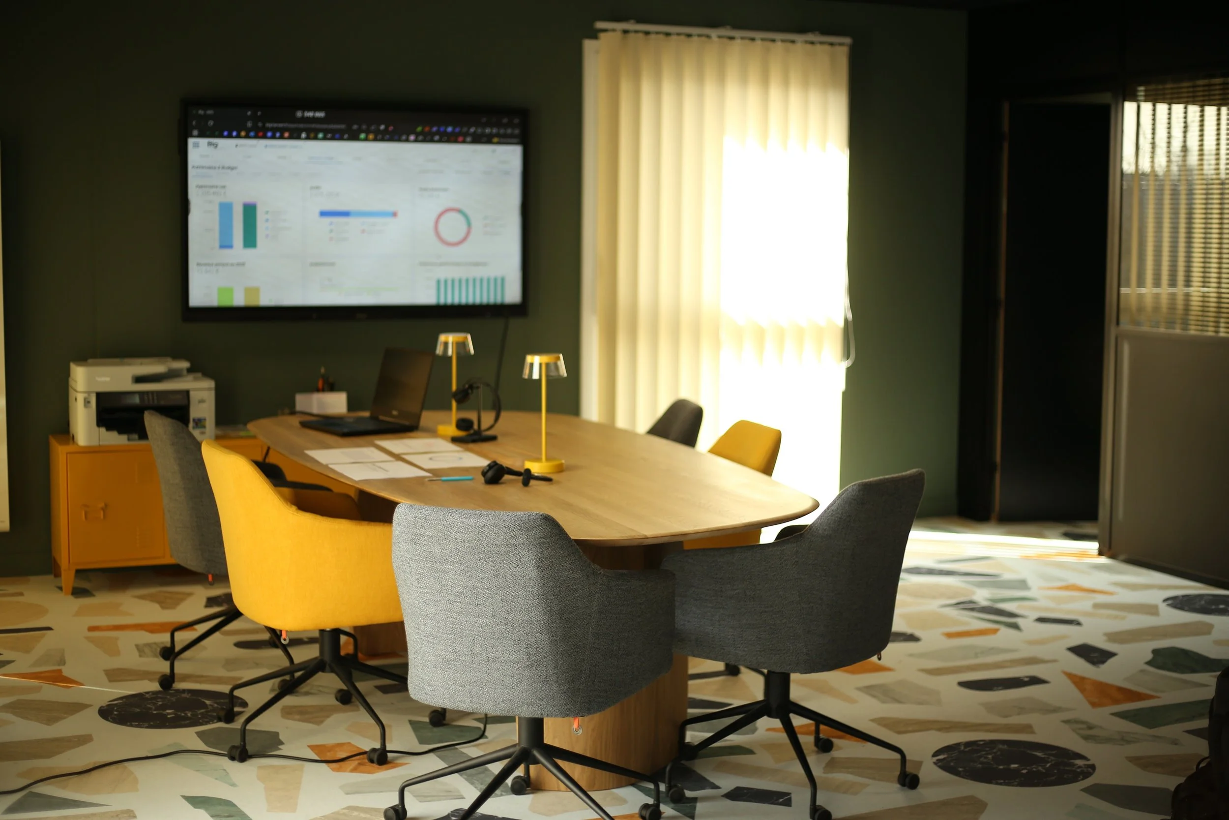 Salle de réunion moderne avec un grand table en bois, six chaises, deux lampes de bureau, un écran à écran plat affichant des graphiques, un ordinateur portable, une imprimante, un fauteuil en jaune, un sol décoratif coloré, et des rideaux blancs laissant passer la lumière naturelle.
