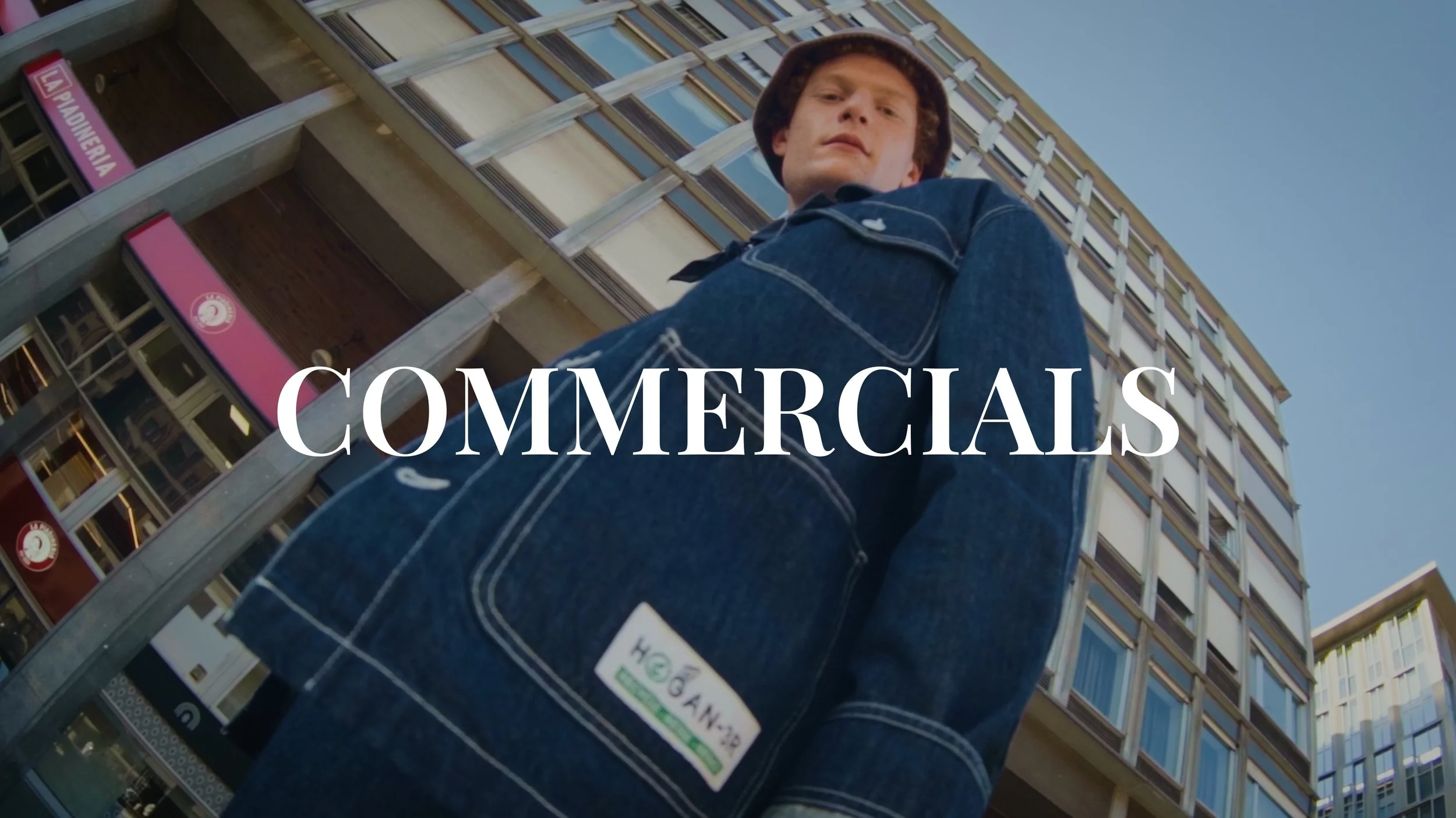 A young man wearing a denim jacket and a bucket hat, looking down at the camera with skyscrapers in the background, with the word 'COMMERCIALS' overlayed on the image.