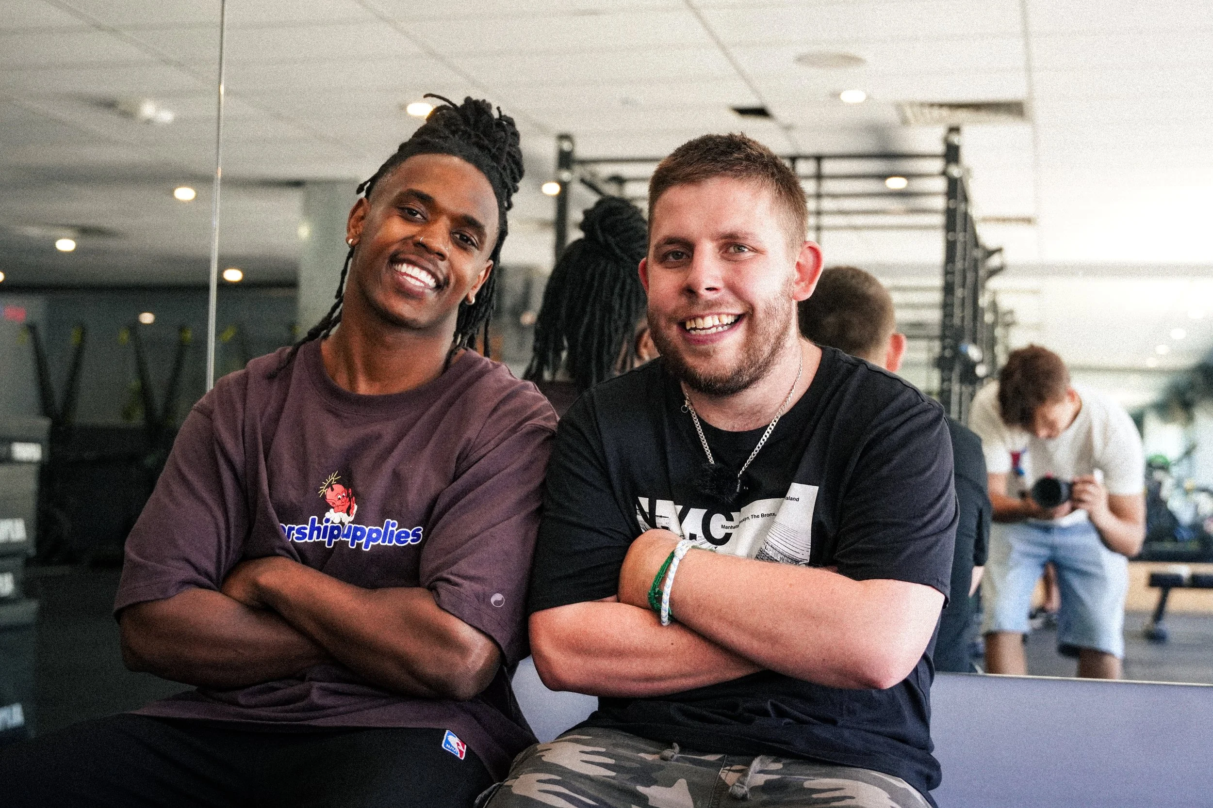 Two smiling men sitting with arms crossed in a gym, with other people working out in the background.