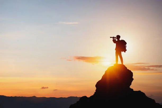 A silhouette of a person standing on a rock, looking through a telescope during a sunset or sunrise.