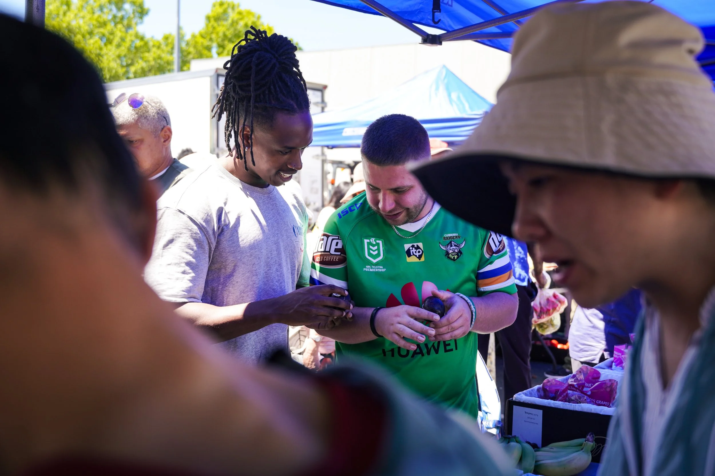 People gathered around at an outdoor market or fair, looking at items and engaging in conversation, with tents and trees visible in the background.