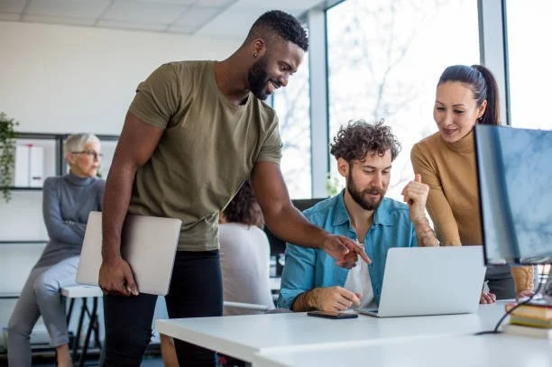 Three diverse coworkers collaborating at a desk with a laptop, in a modern office setting, with other employees working in the background.