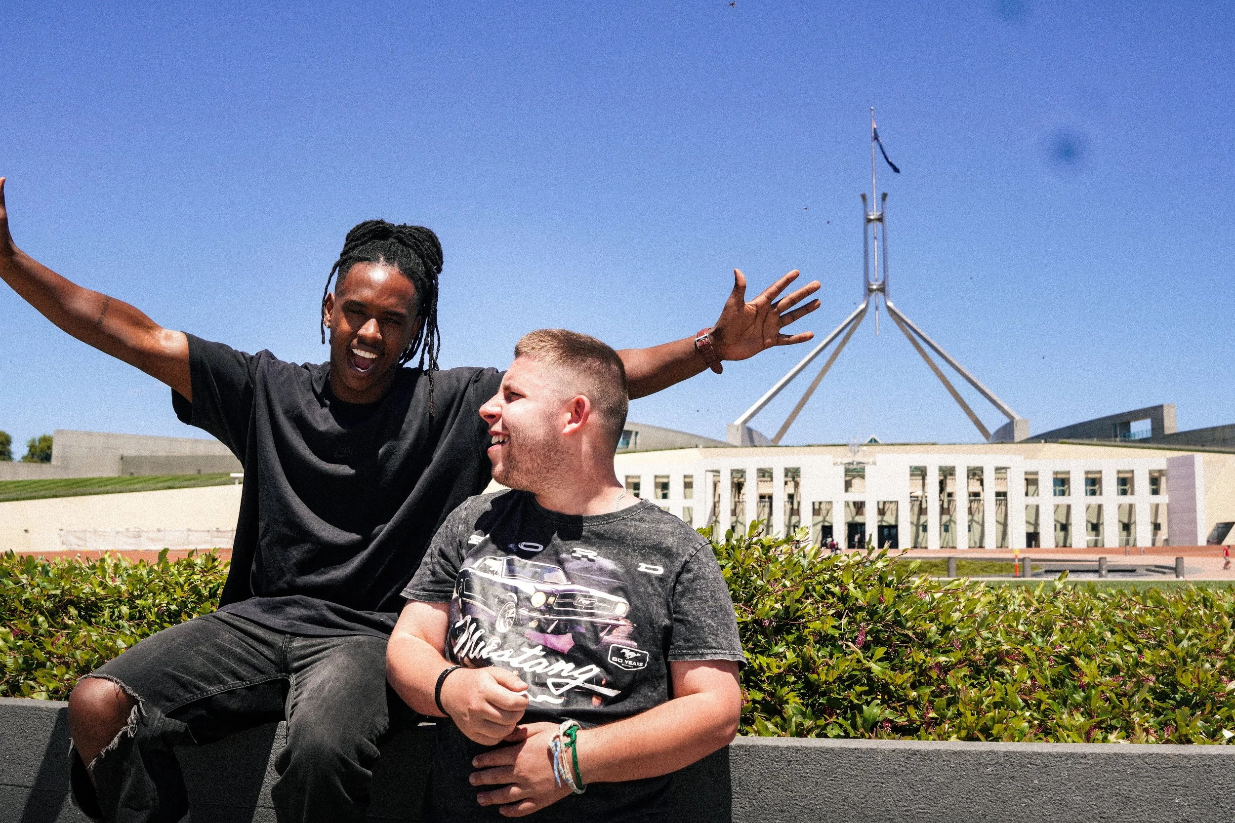 Two men sitting outdoors in front of the Australian Parliament House, one with arms raised and both smiling.