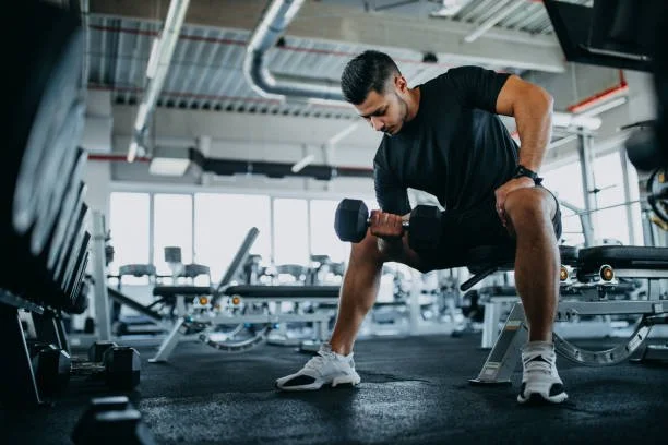 A man exercising with a dumbbell in a gym setting, wearing a black shirt and shorts.