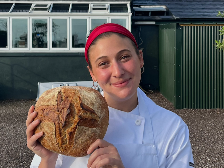 Chef Chandler Maizel in a white chef's coat and red headband holding a large sourdough loaf with a carved cross on top, standing outdoors in front of a modern building with glass windows and a green fence.