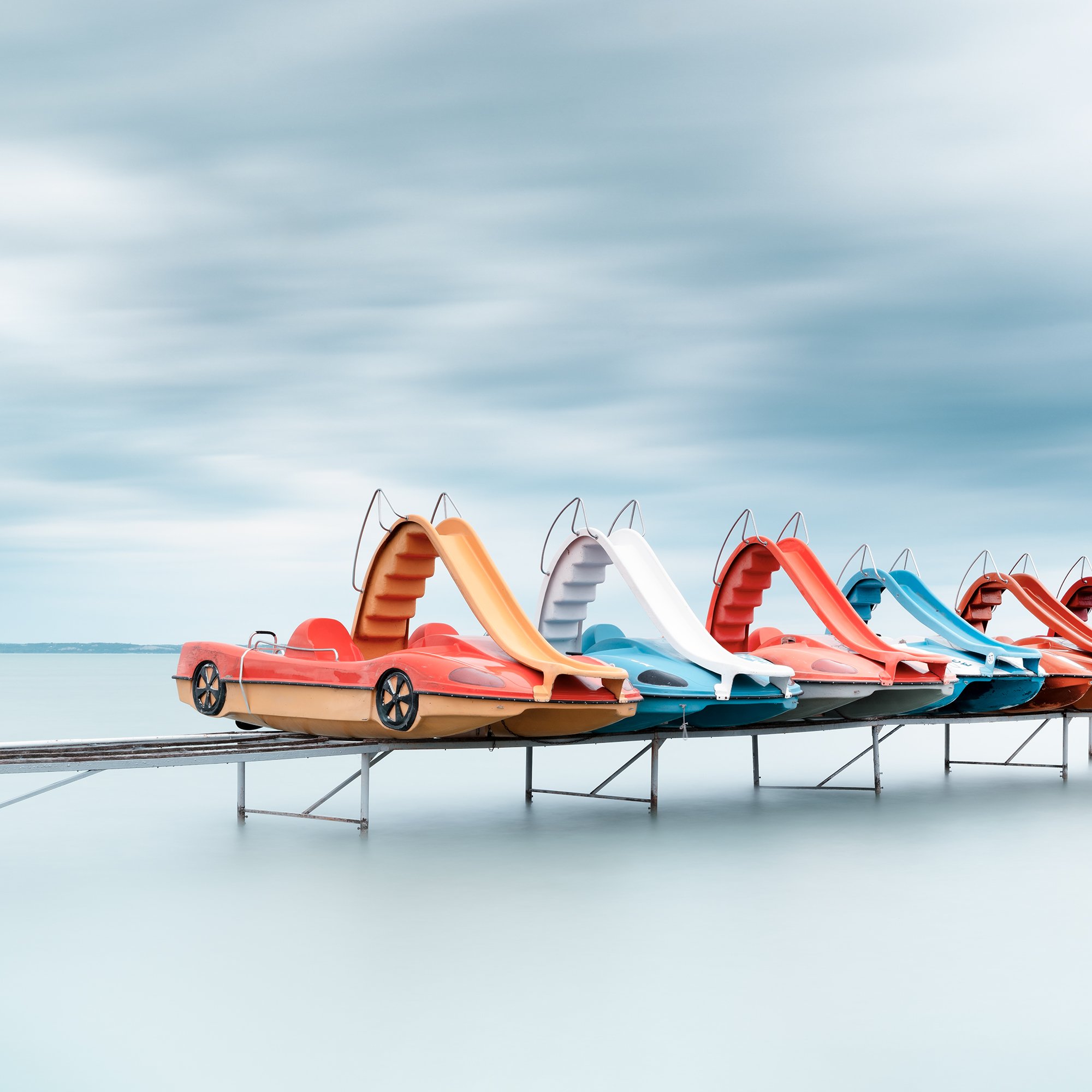 Gerald Berghammer - Color waterscape photography. A row of colorful plastic pedal boats shaped like race cars on a dock over water, under a cloudy sky. Print detail 1