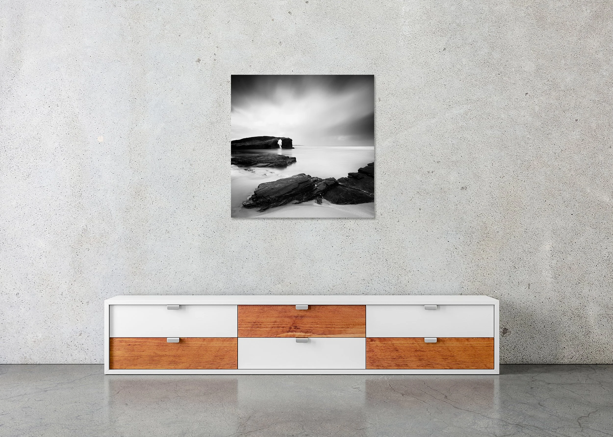 Frameless ChromaLuxe metal print showing a monochrome coastal scene with a natural rock arch, rocky beach and smooth water under an overcast sky.