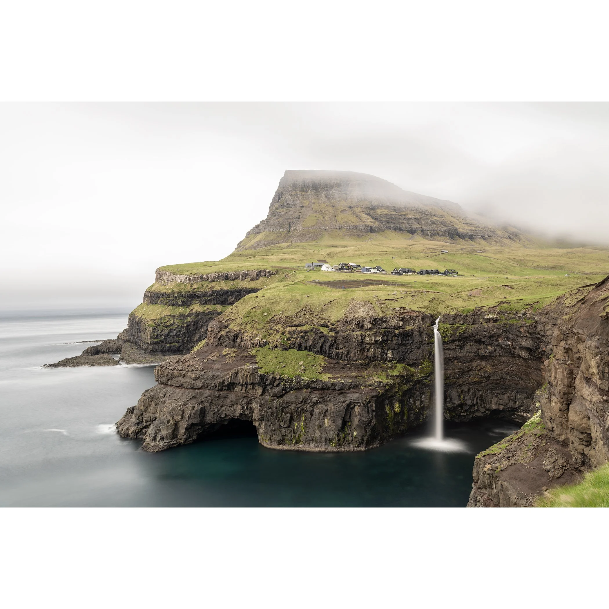 Gerald Berghammer - Color Photography. Coastal landscape featuring a high plateau with a waterfall cascading into the ocean, partially obscured by fog.