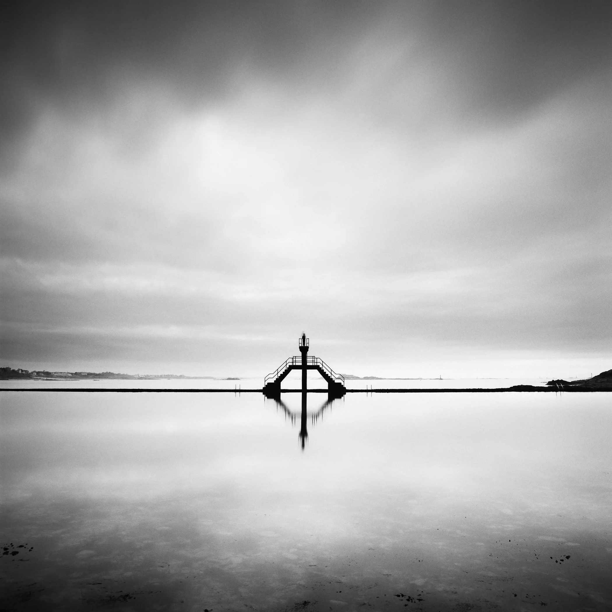 Black-and-white minimalist seascape with a symmetrical diving platform reflected in calm water beneath a cloudy sky.