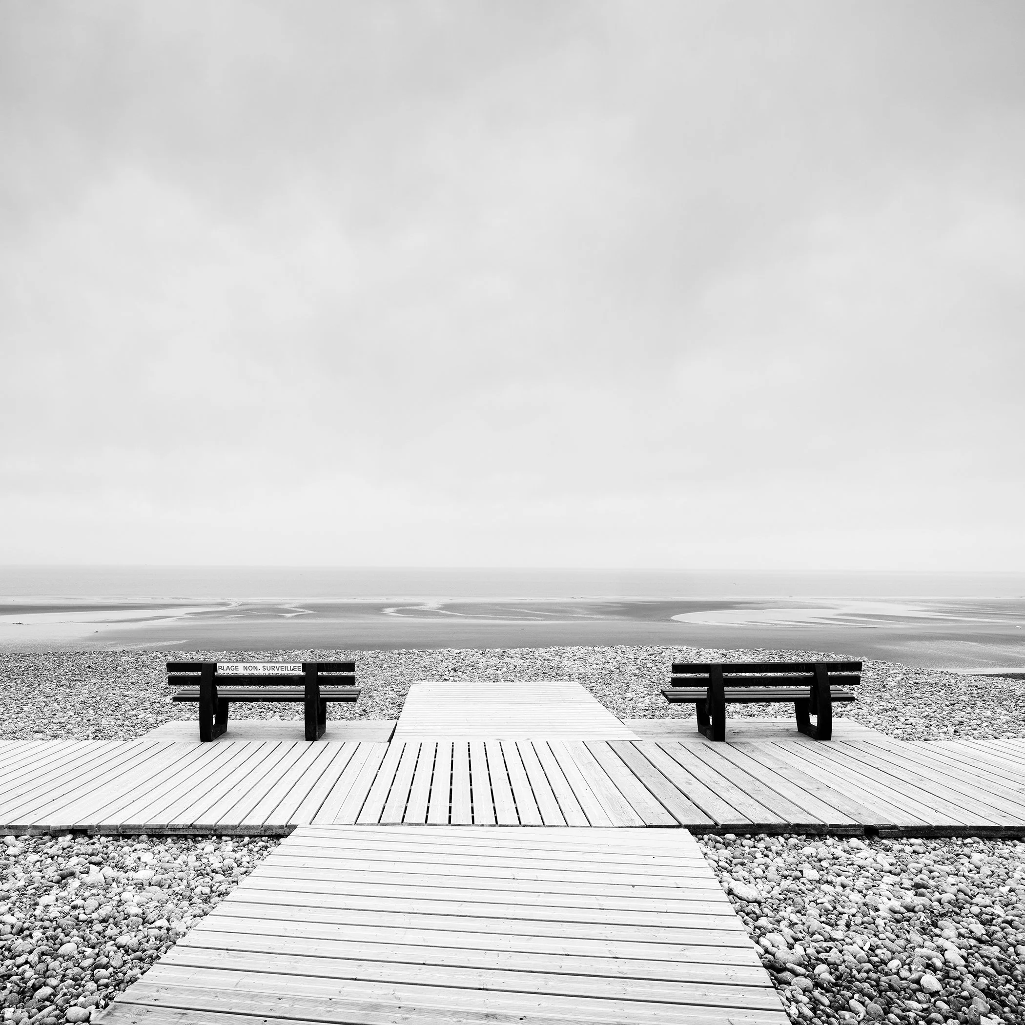 Black-and-white minimalist landscape photo of benches facing a quiet, deserted French beach, with soft light and a wide horizon.