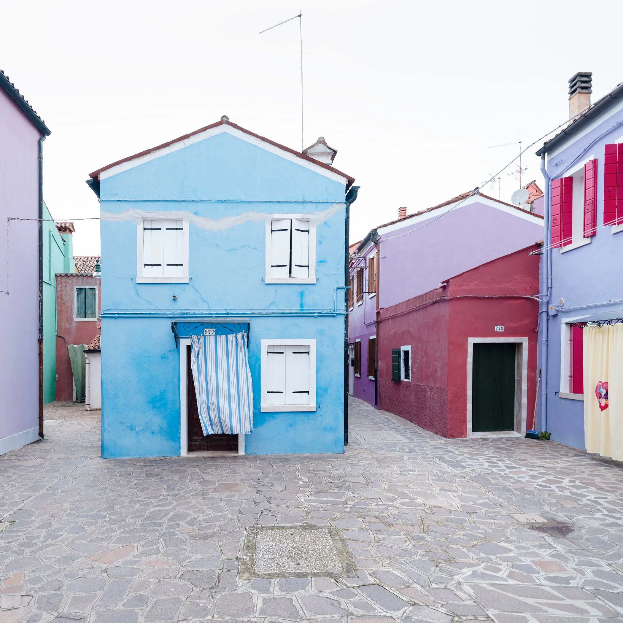 © 2025 Gerald Berghammer photo - Colorful houses in pastel shades on a cobblestone street, with colorful windows and a striped curtain hanging in front of one house.