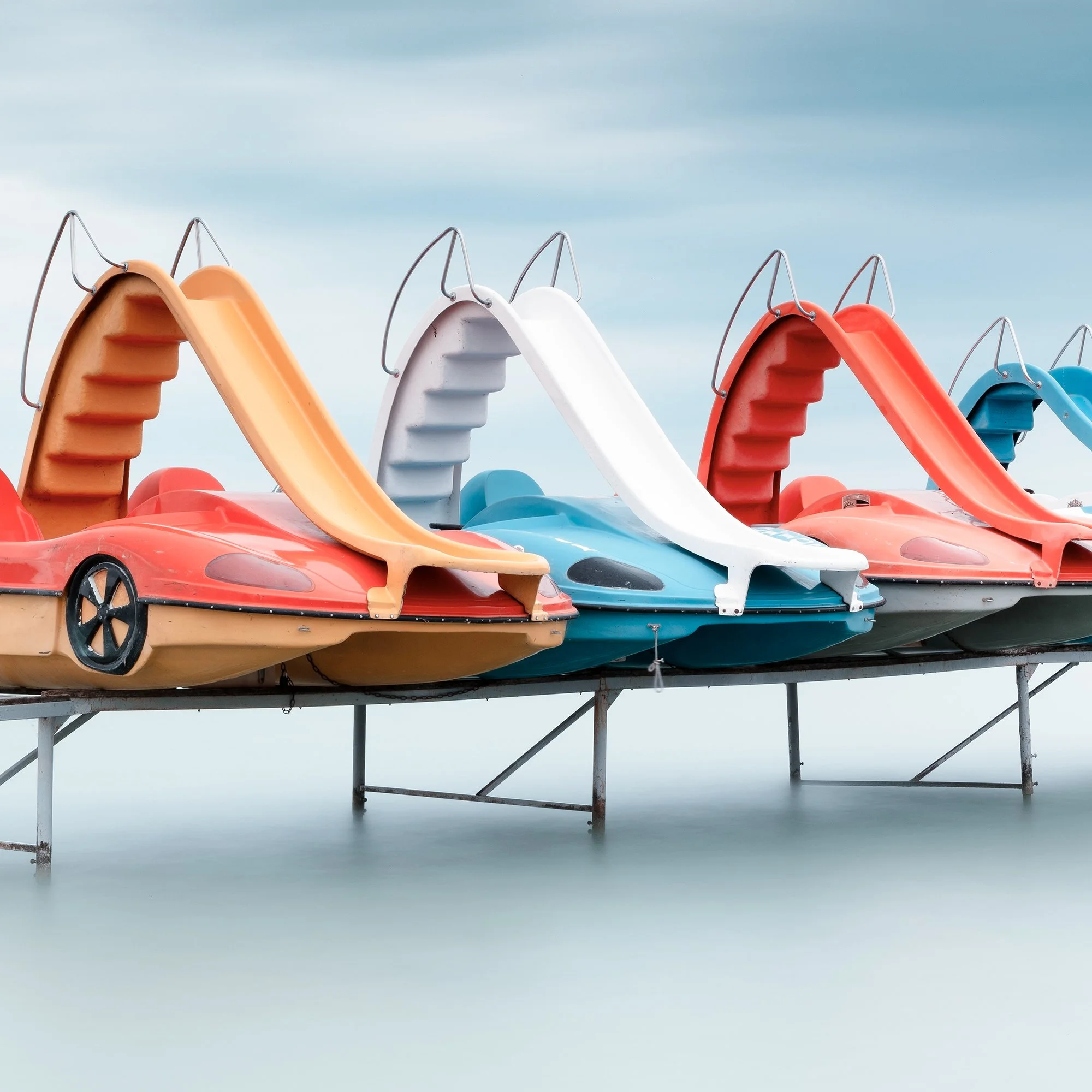 Gerald Berghammer - Color waterscape photography. A row of colorful plastic pedal boats shaped like race cars on a dock over water, under a cloudy sky. Print detail 3