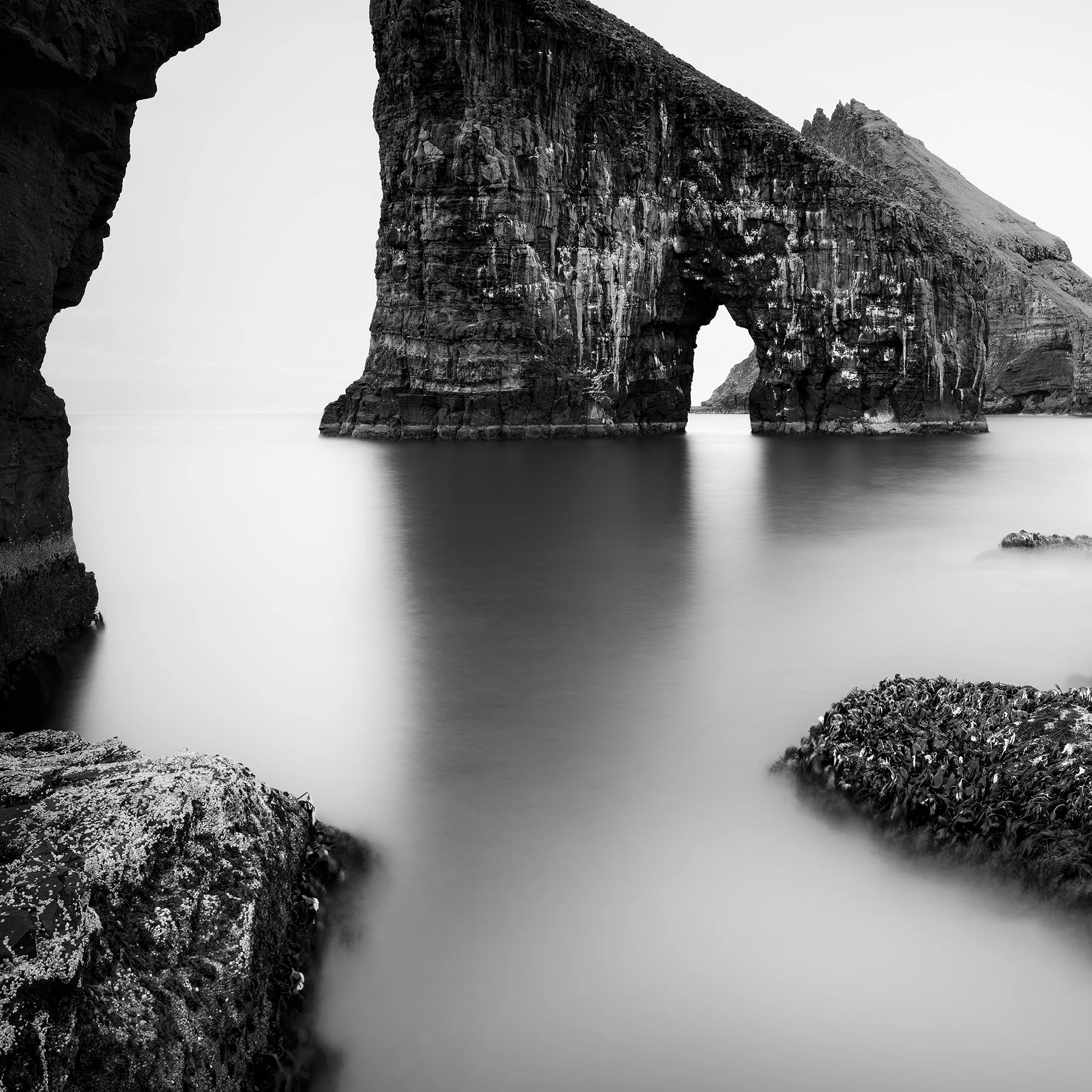 Black and white coastal landscape with a striking sea stack arch and soft long-exposure water – close-up