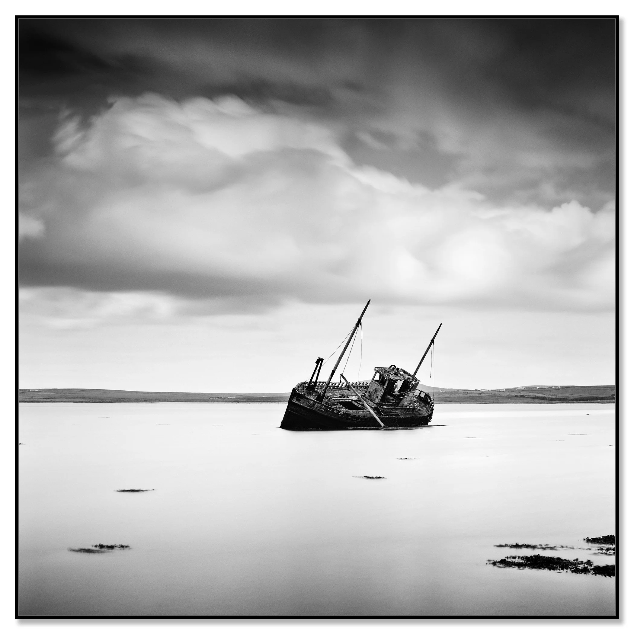 Black-and-white photo of a shipwreck in shallow water under a cloudy sky, distant shoreline on the horizon – framed ArtBox black