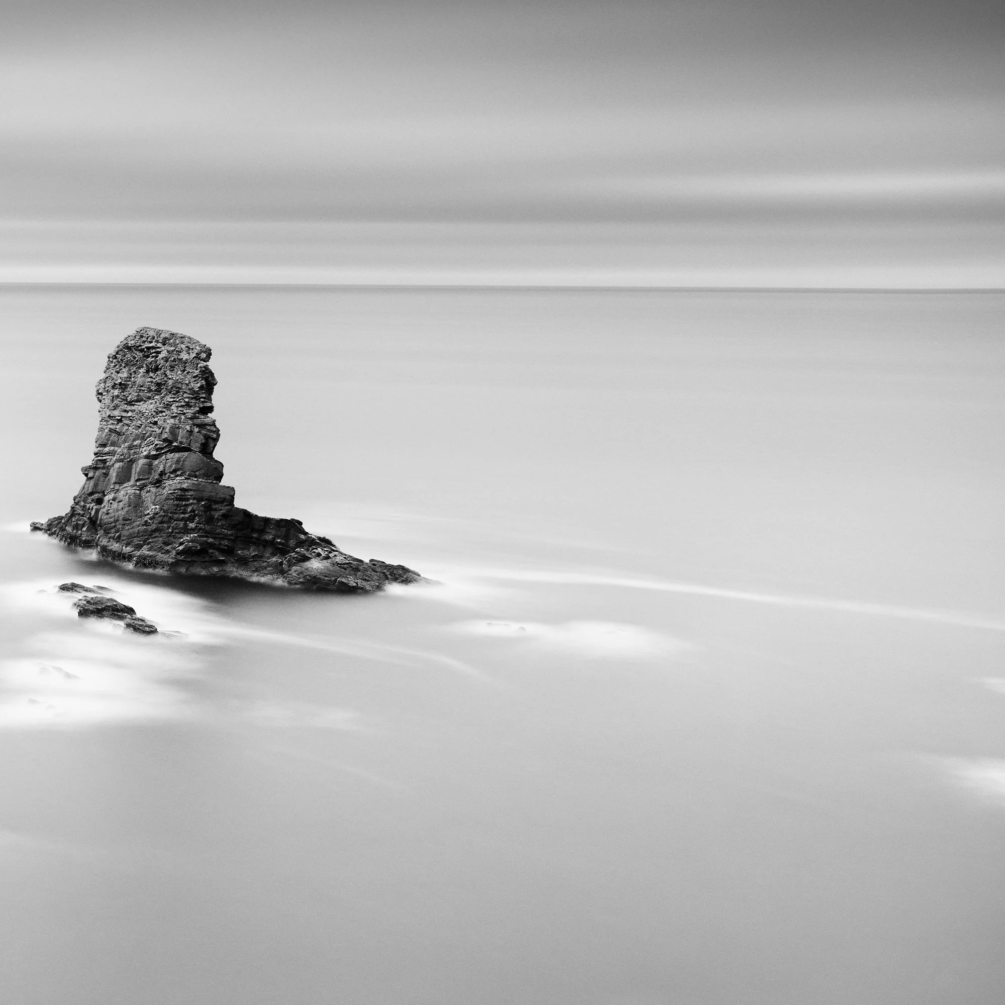 Gerald Berghammer - Black and white panorama seascape photography. Rocky coastline with cliffs and a tall rock formation in the water, under a cloudy sky. Print detail 2