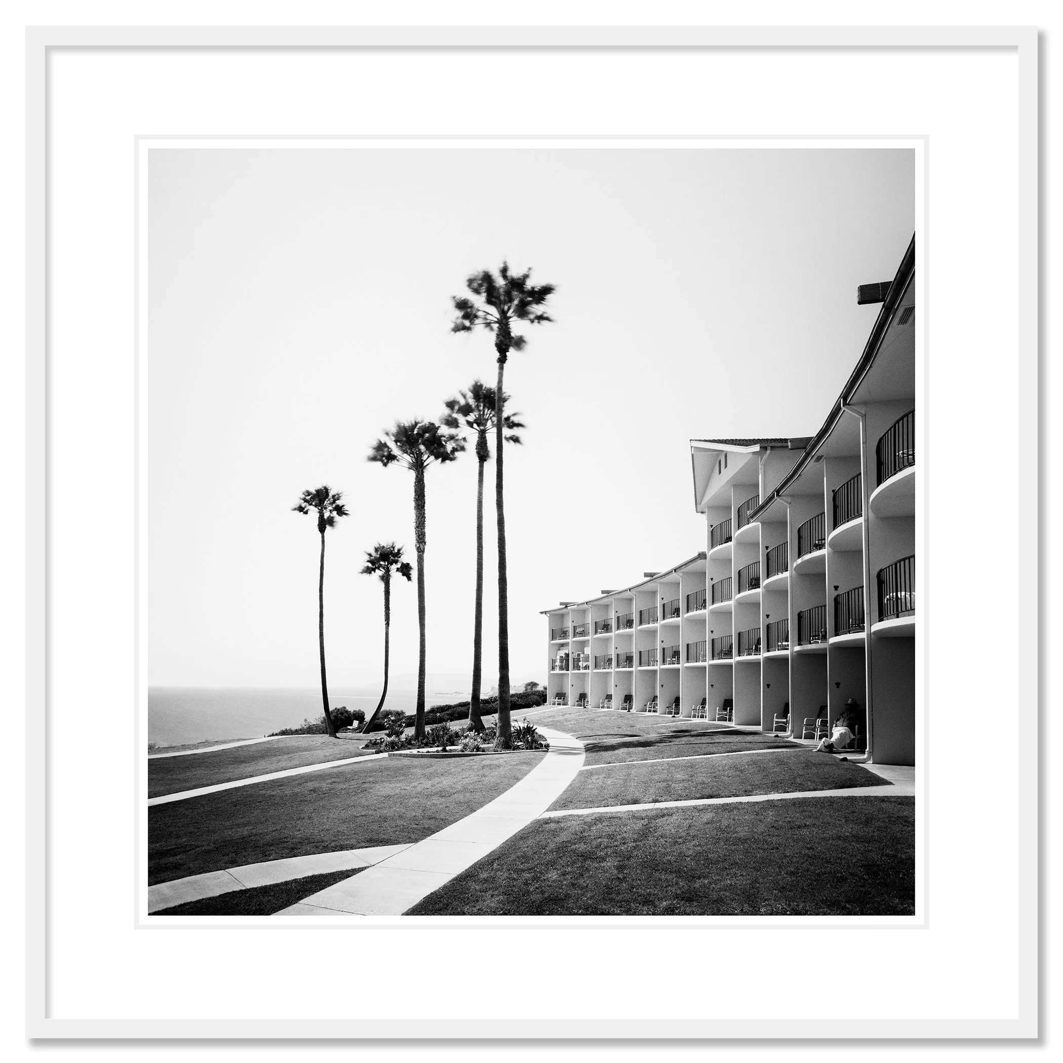 Gerald Berghammer - Black and white landscape photography. Seaside hotel with multiple balconies, a curved walkway, and tall palm trees near the ocean. Classic framed white