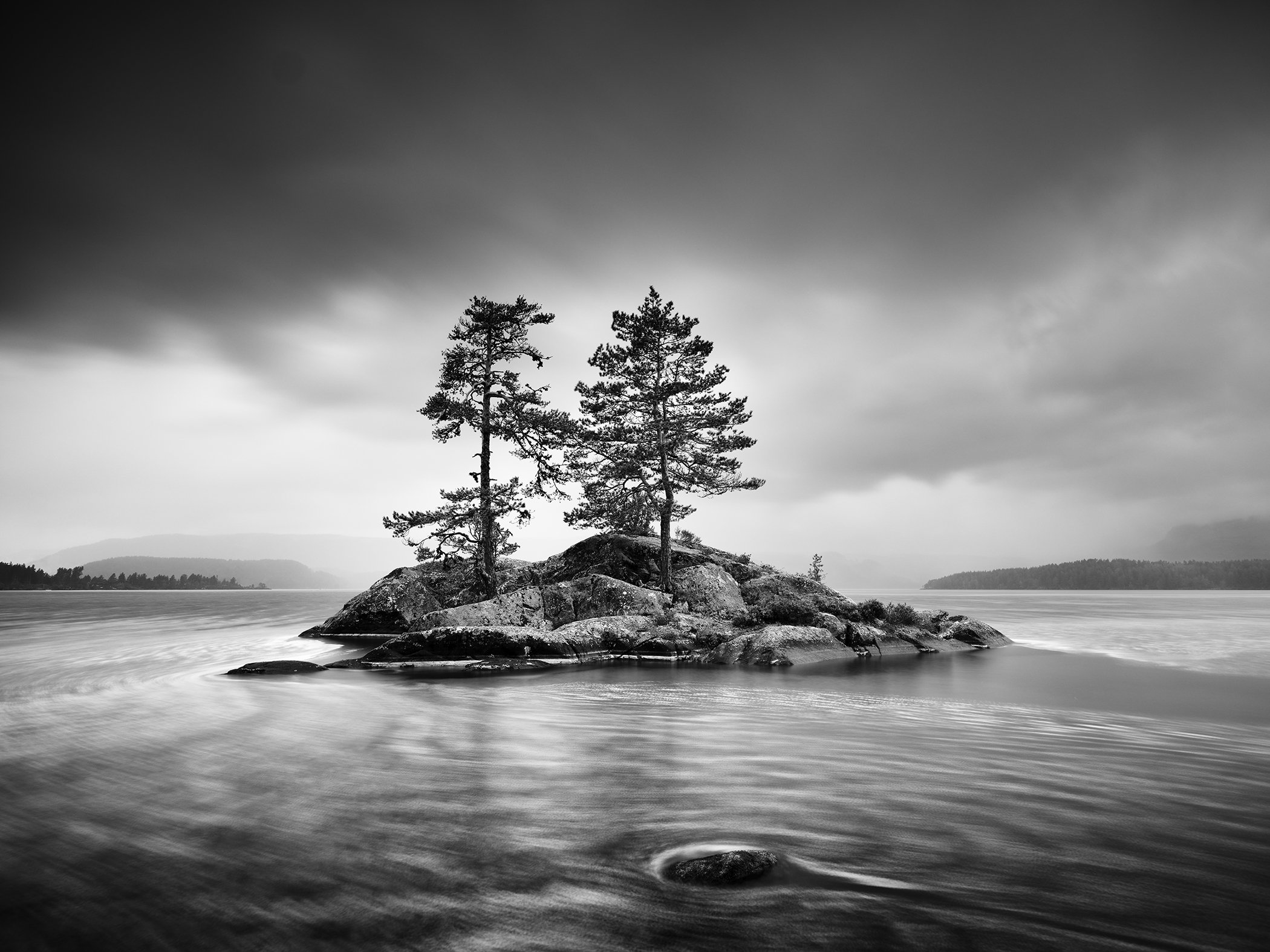 Monochrome landscape photograph showing pine trees on a rocky island surrounded by calm water and dramatic clouds, by Gerald Berghammer.