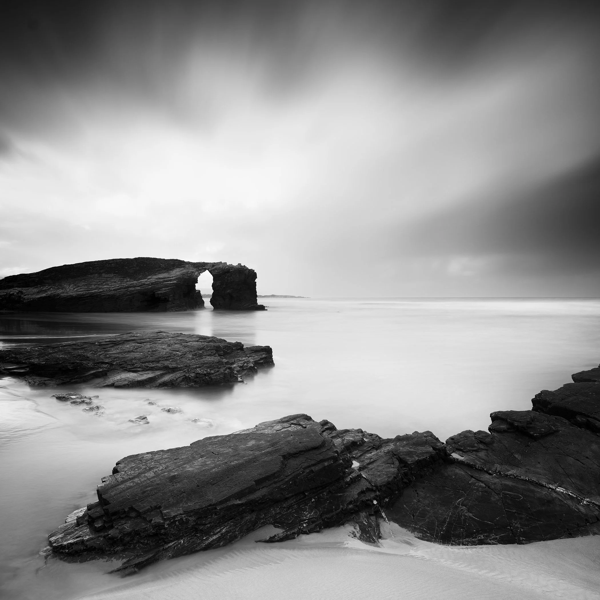 Black-and-white seascape with rock arch on a rocky shoreline and calm water under a cloudy sky.