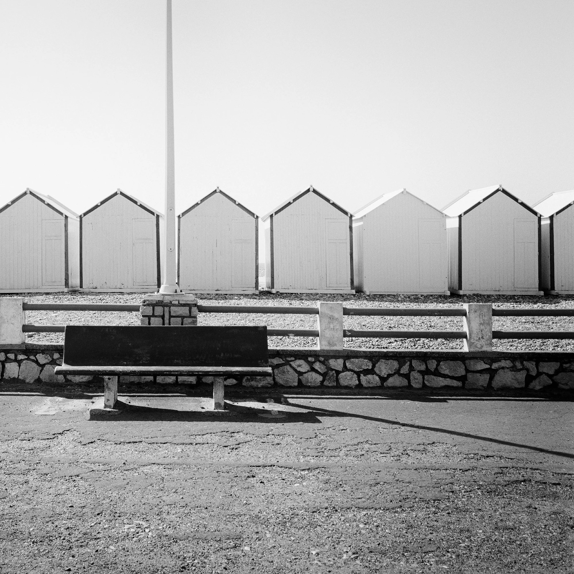 Empty bench on the promenade facing beach huts, centred streetlamp, minimalist black-and-white coastal photograph, Detail 2
