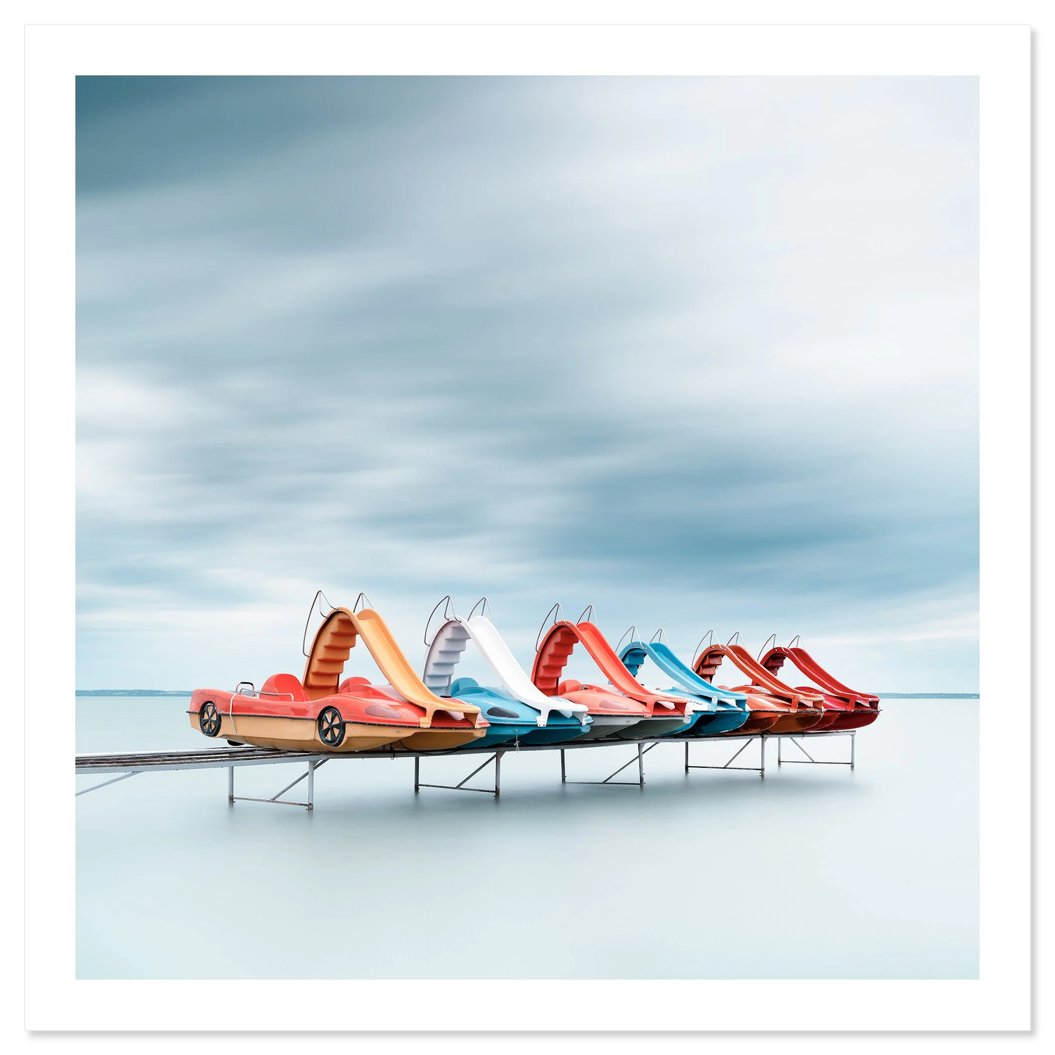 Gerald Berghammer - Color waterscape photography. A row of colorful plastic pedal boats shaped like race cars on a dock over water, under a cloudy sky. Fine art print only