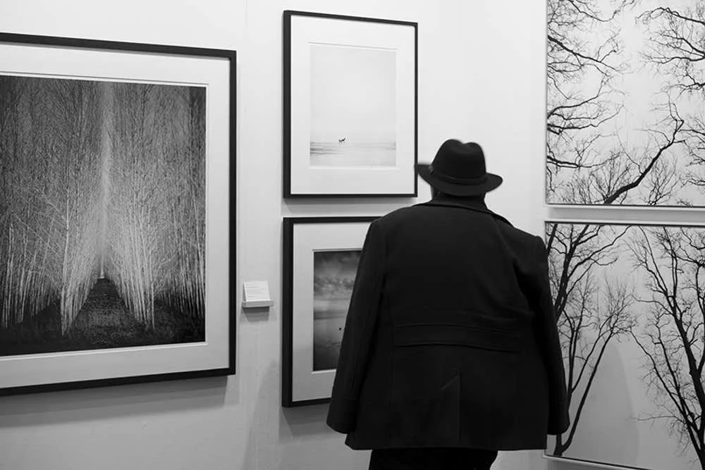 A visitor wearing a dark coat and hat views framed black-and-white photographs displayed on a white gallery wall at art Karlsruhe, presented by Silverfineart