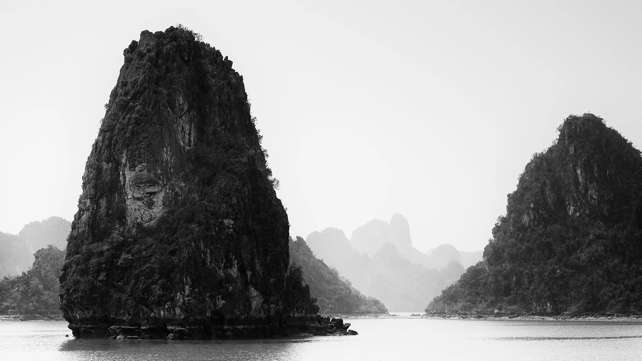© 2025 Gerald Berghammer - Black and white landscape photograph of limestone karst islands in Ha Long Bay, Vietnam, surrounded by calm water and distant misty peaks