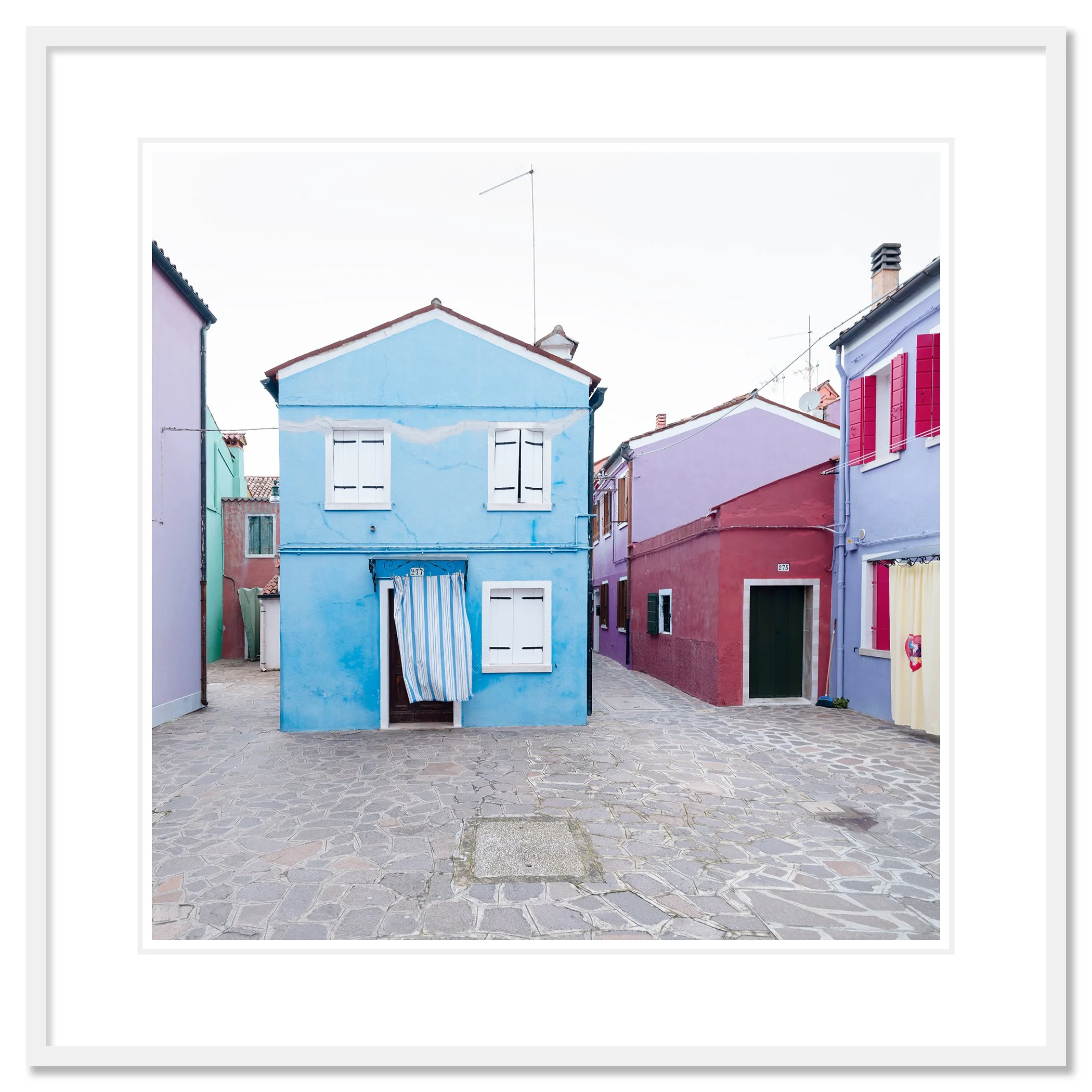 © 2025 Gerald Berghammer photo - Colorful houses in pastel shades on a cobblestone street, with colorful windows and a striped curtain hanging in front of one house. Classic framed white