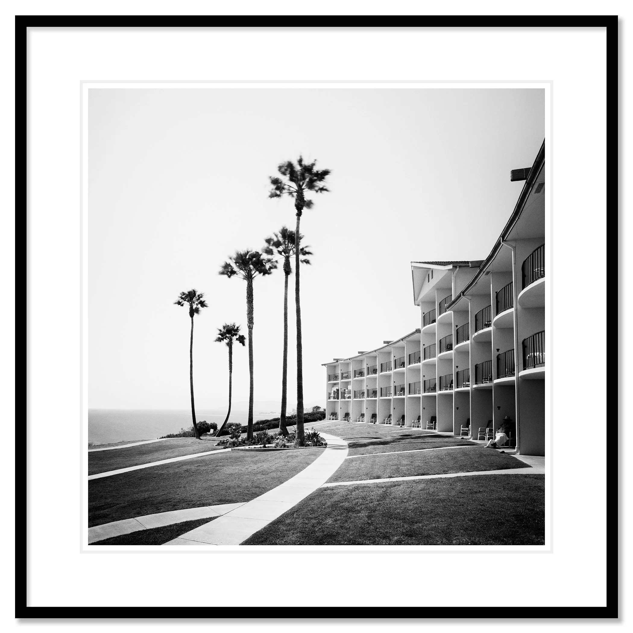 Gerald Berghammer - Black and white landscape photography. Seaside hotel with multiple balconies, a curved walkway, and tall palm trees near the ocean. Classic framed black