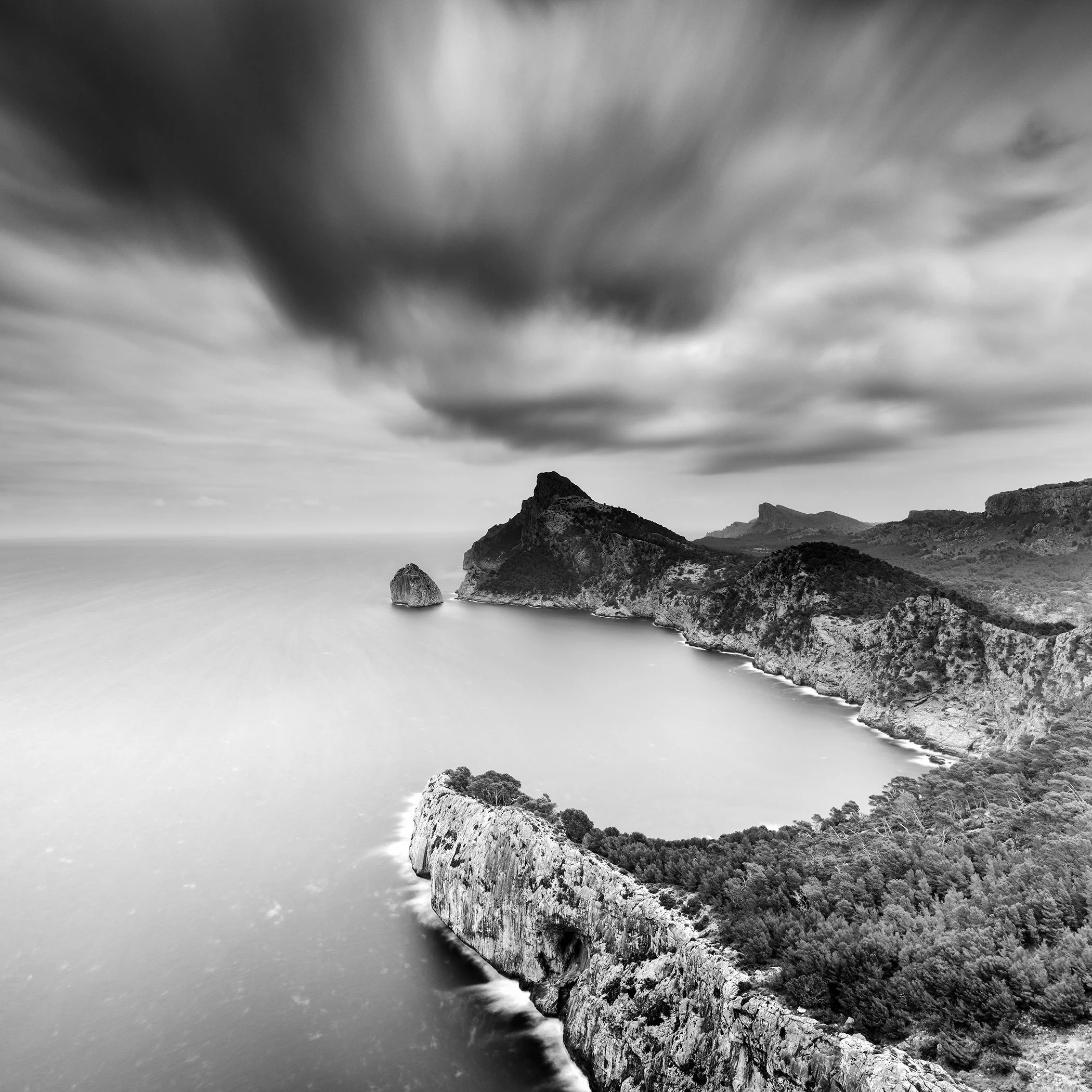 Black-and-white long-exposure photo of rugged coastal cliffs around a calm bay, with smooth water and dramatic grey clouds over the sea.