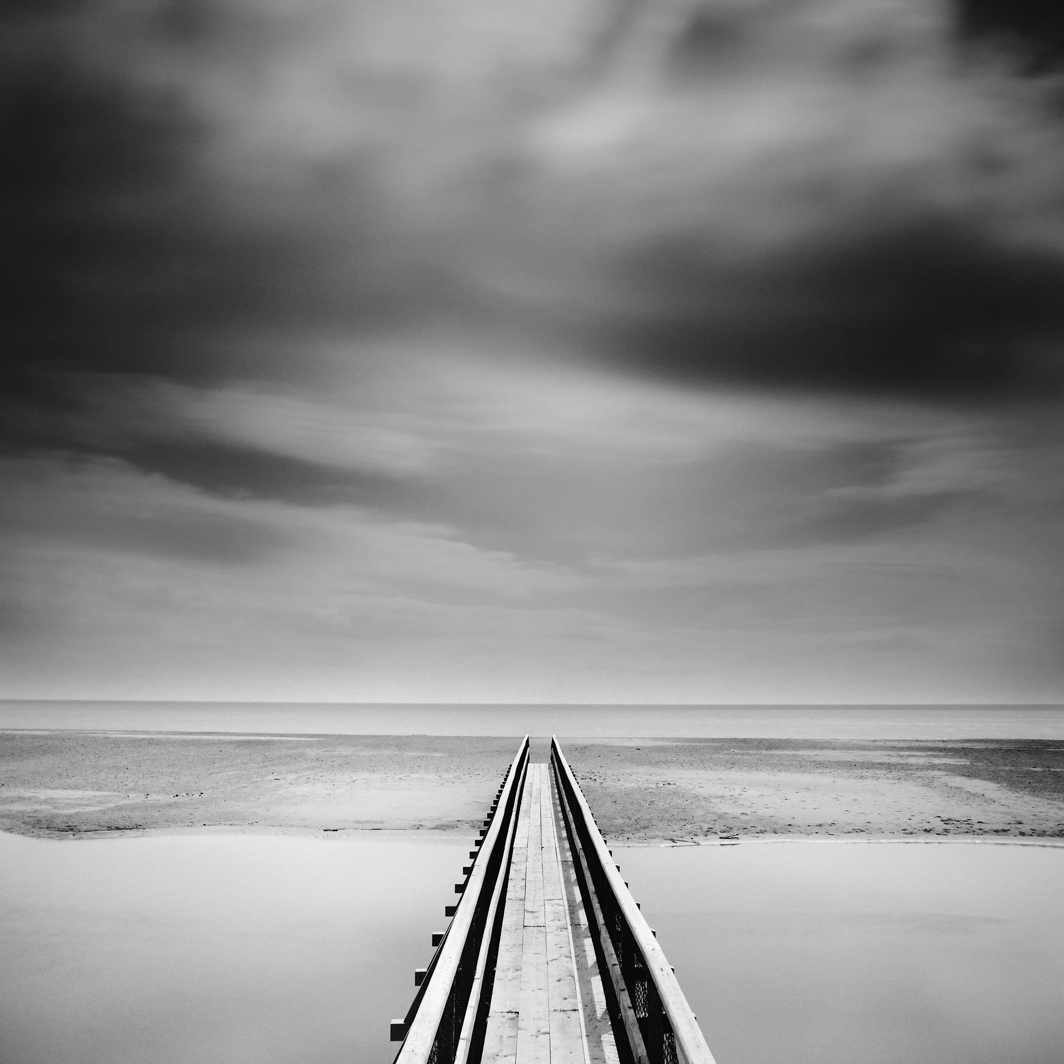 Black-and-white photograph of a centred wooden pier crossing calm water towards a sandy beach and open sea in Ireland by Gerald Berghammer.