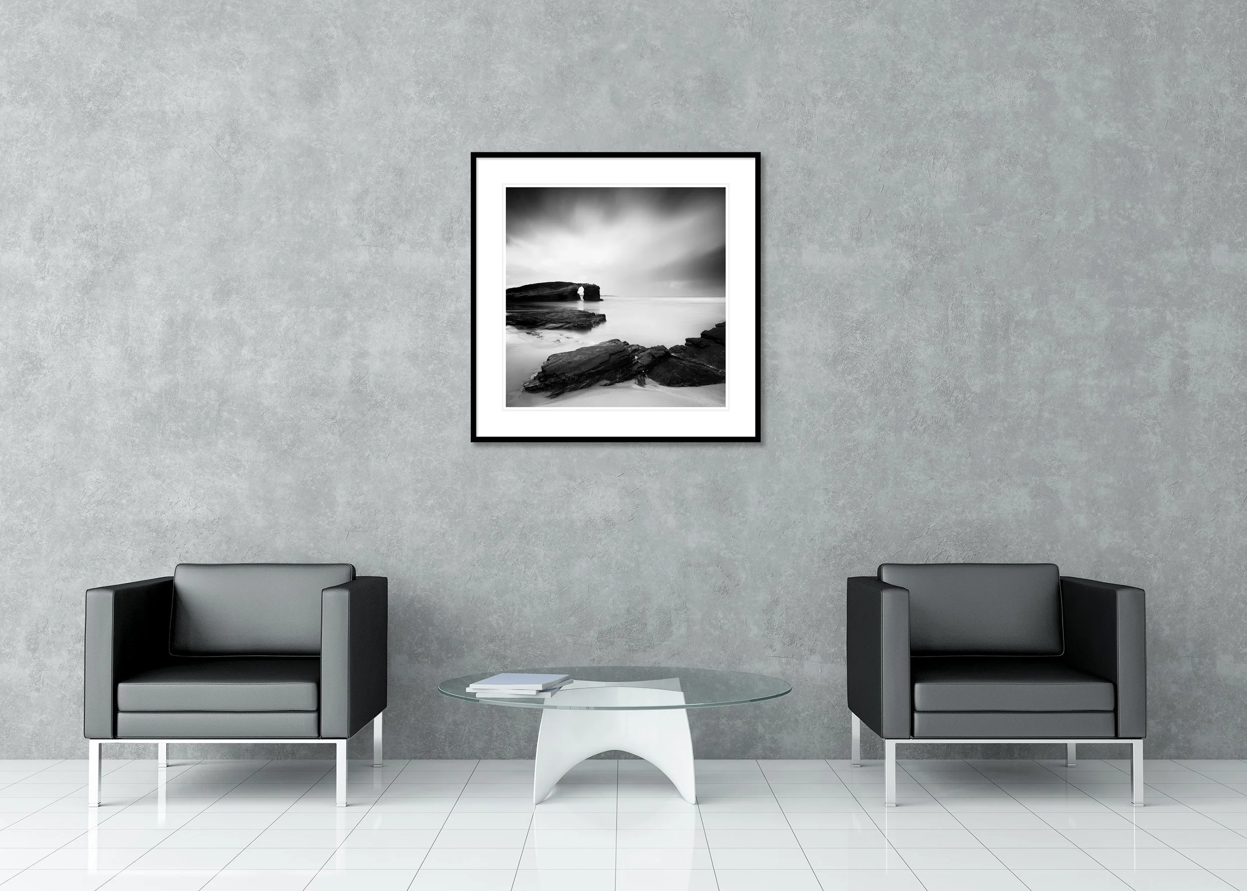Monochrome coastal photograph of a natural rock arch and rocky beach with smooth water under an overcast sky, archival pigment print in a classic frame with a mount.