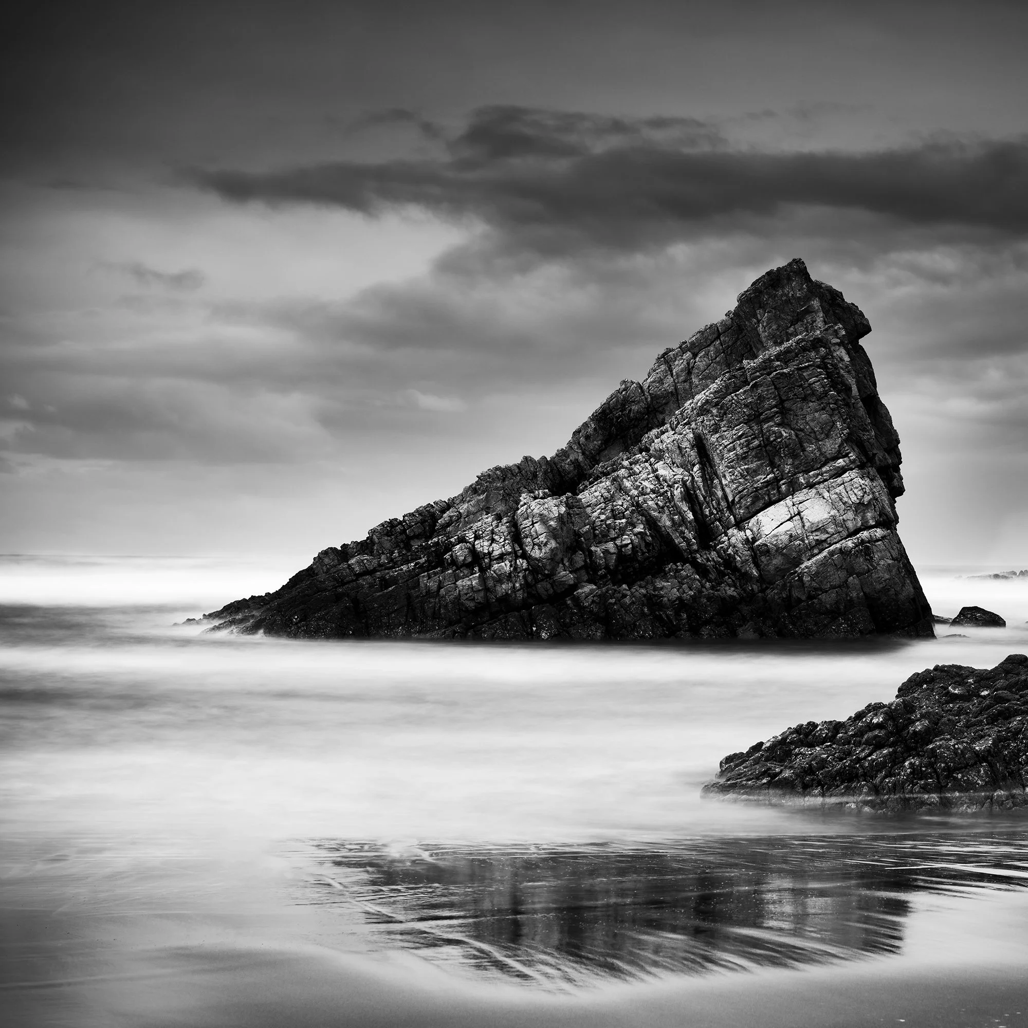© 2023 Gerald Berghammer. Black-and-white photograph of jagged sea rocks emerging from calm water under a cloudy sky. Print detail 1